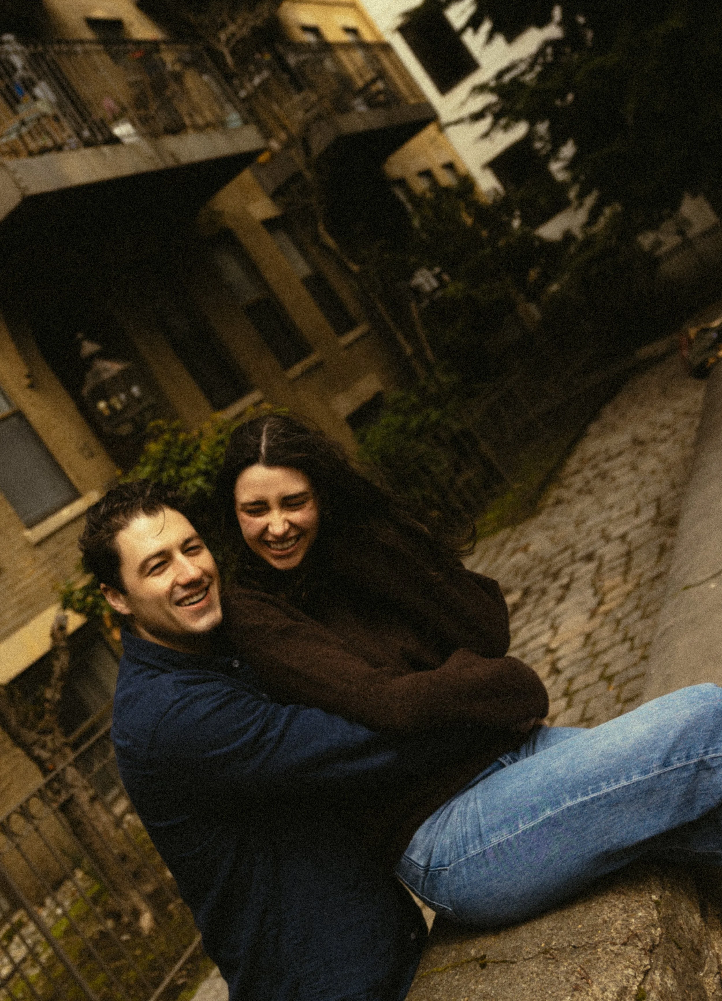 Two people smiling and sitting close together outdoors at night, with a building and trees in the background.