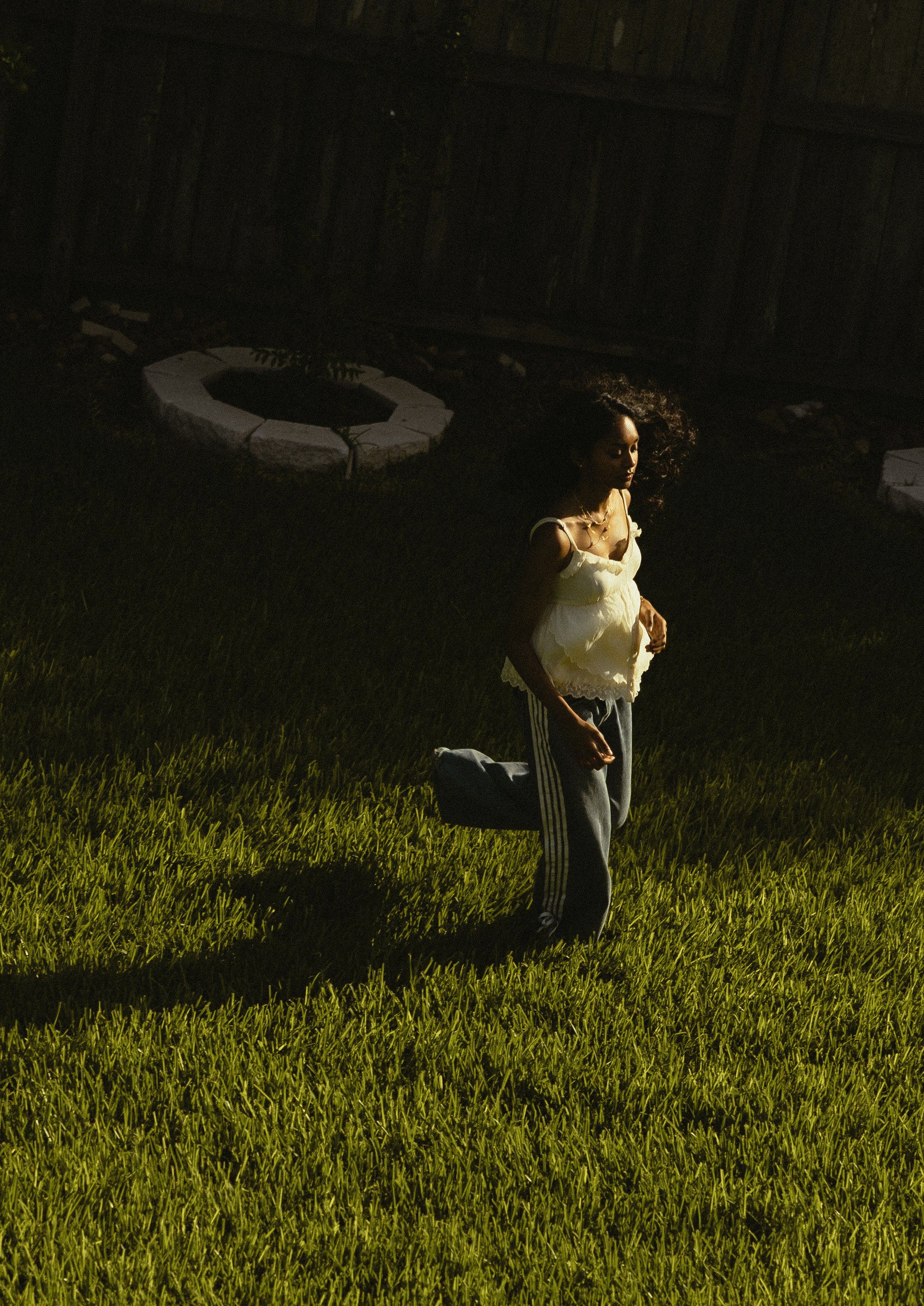 A woman standing outdoors on green grass with dark shadows, wearing a yellow sleeveless top and gray pants with black stripes, looking down with her eyes closed and curly hair blowing in the wind.