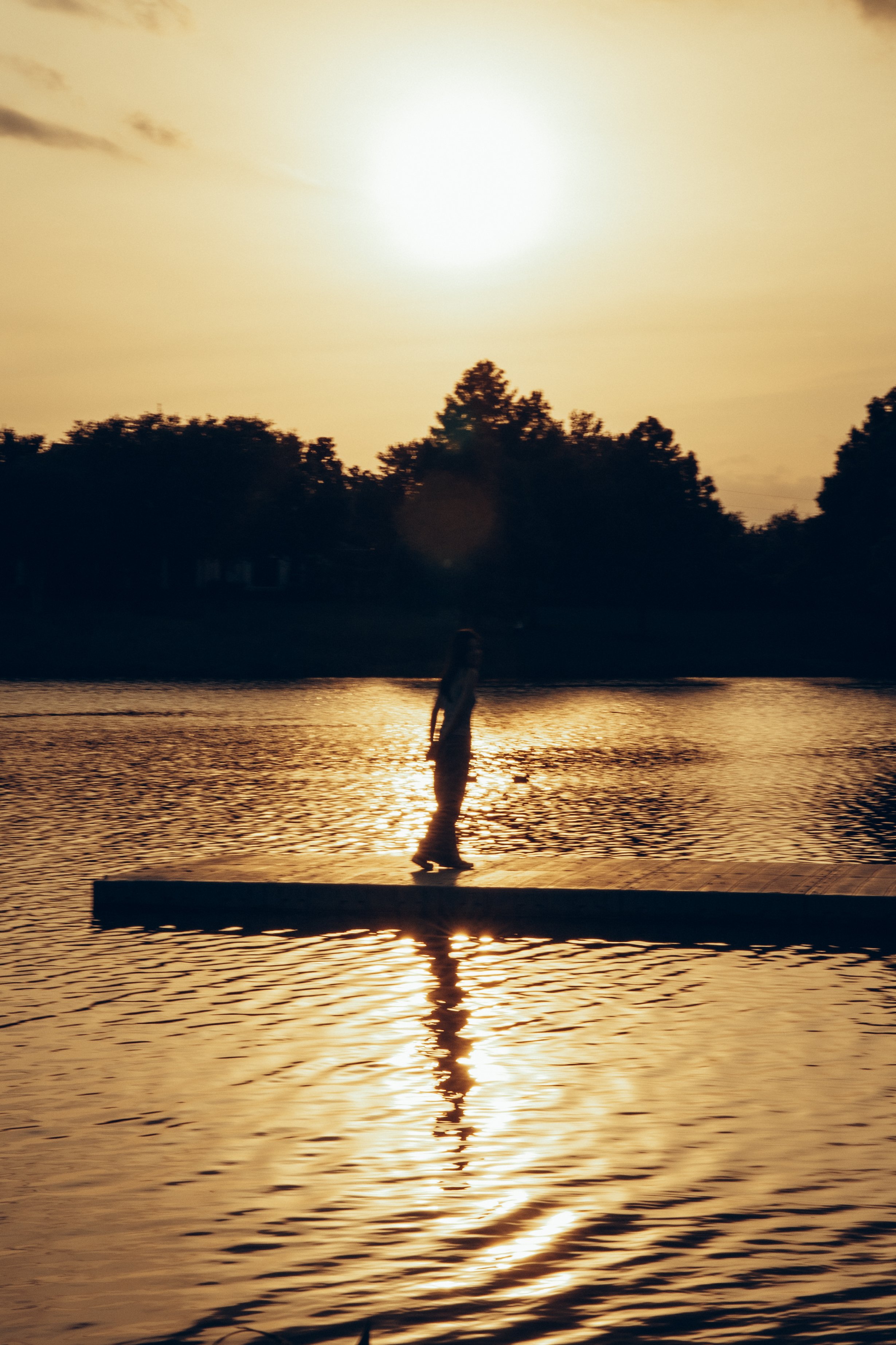 Person walking on a dock by a body of water during sunset, with trees in the background and reflections on the water.