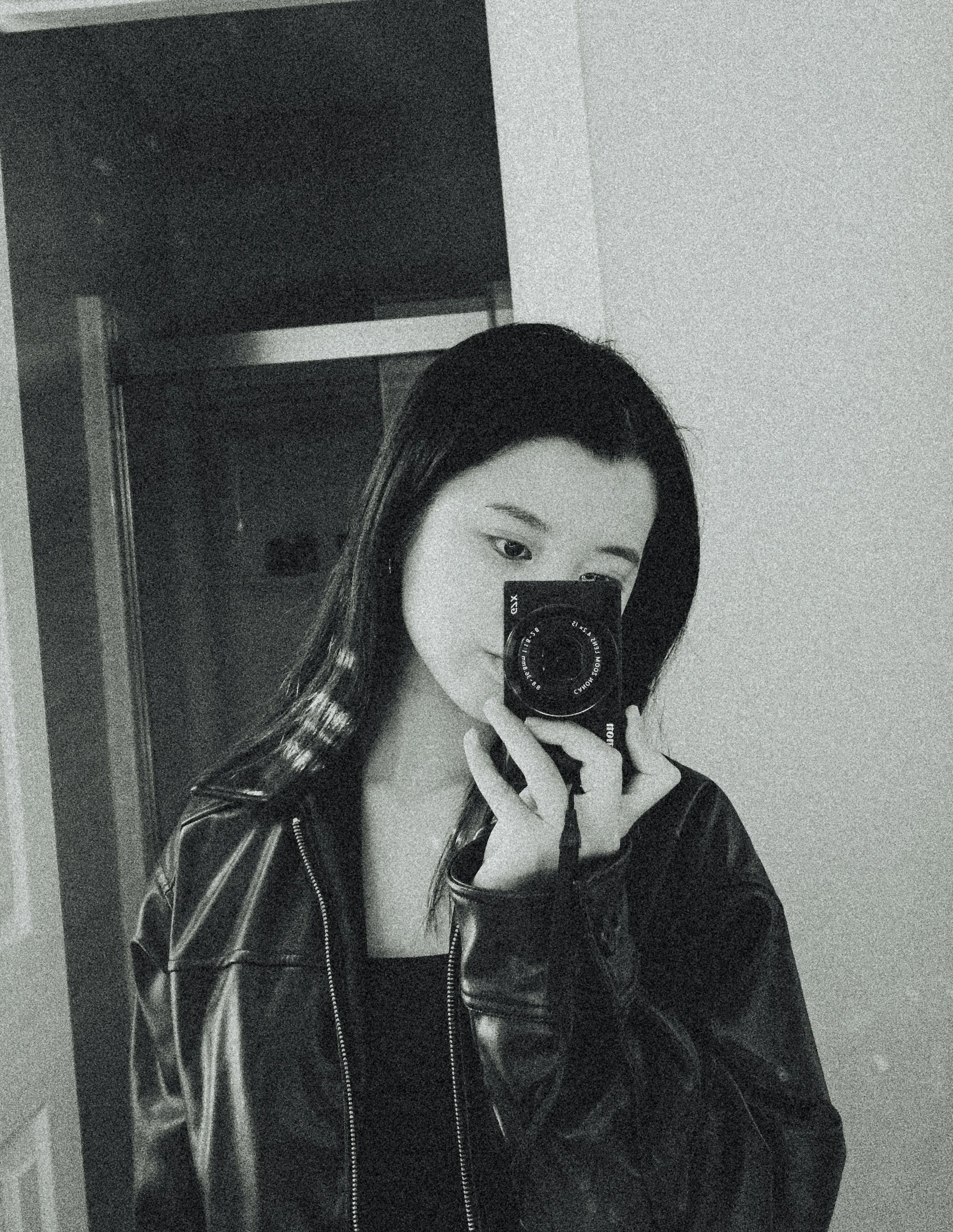 Catherine with dark hair taking a mirror selfie with a camera, wearing a black leather jacket and a black top.