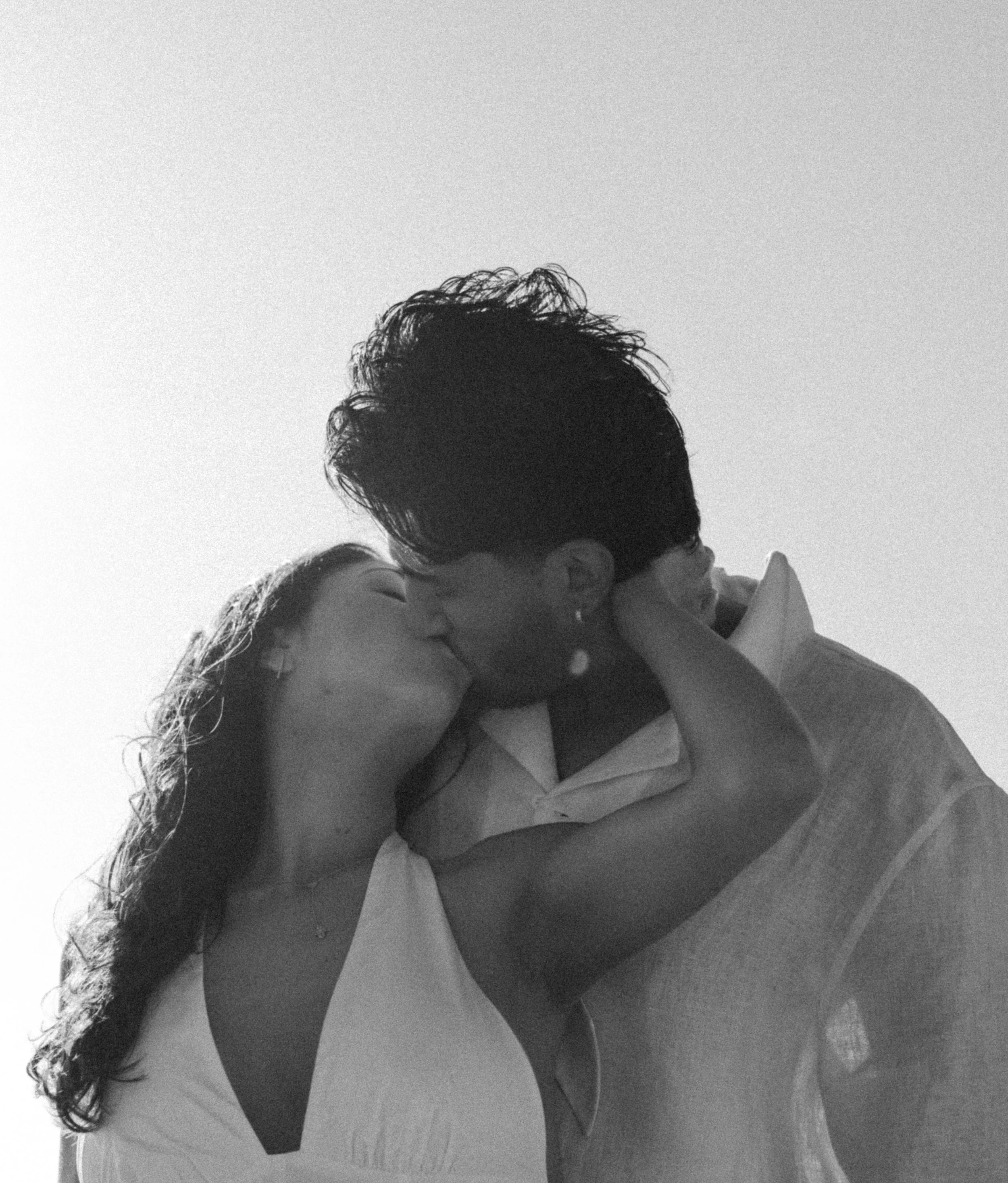 A black and white photograph shows a couple embracing and kissing on a bed. The woman with long hair is lying on her back with her arms around the man. The man with curly hair is leaning over her in a tender moment.