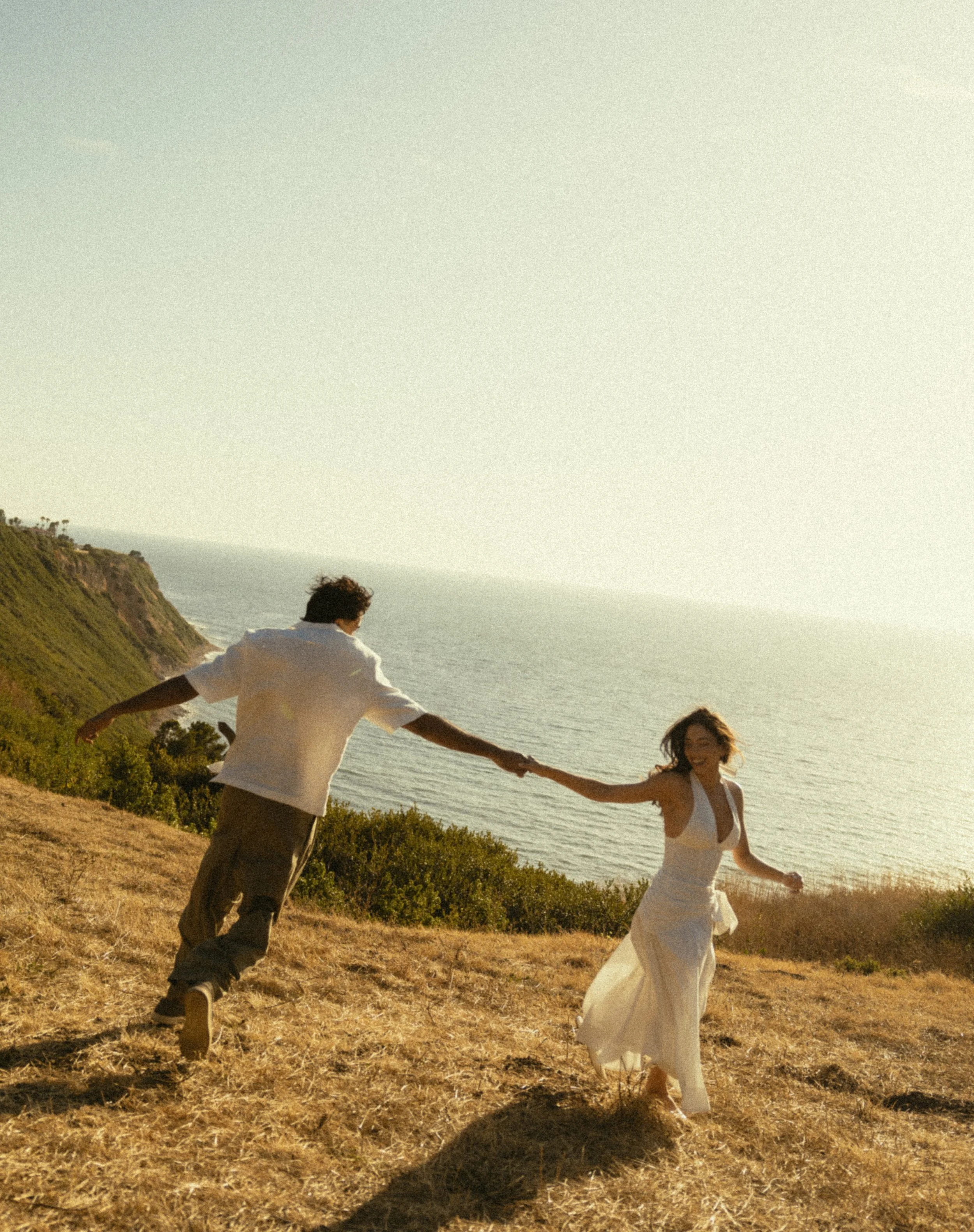 A couple holding hands and walking on a grassy hillside near the coast during sunset, with the ocean in the background.