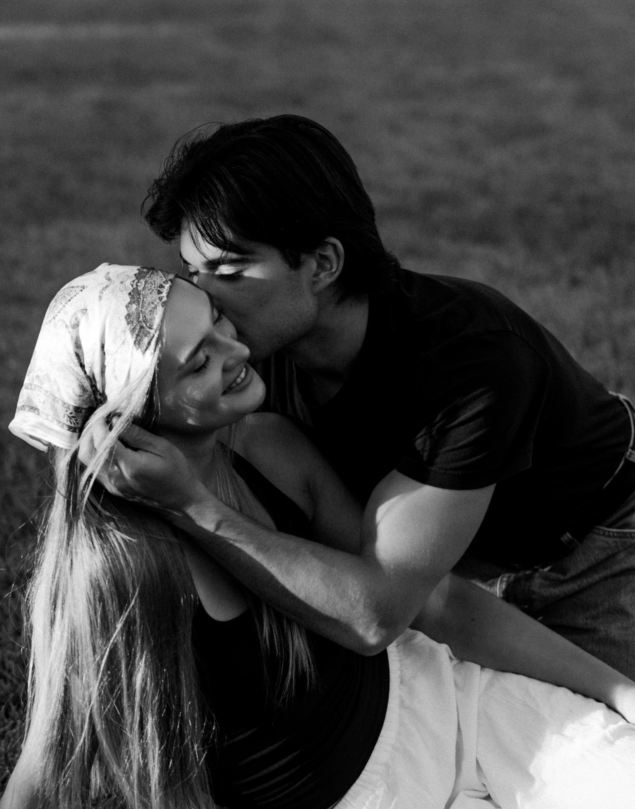 A black and white photo of a man and woman embracing each other closely, with the man kissing the woman's forehead and their eyes closed, outdoors with blurred background.