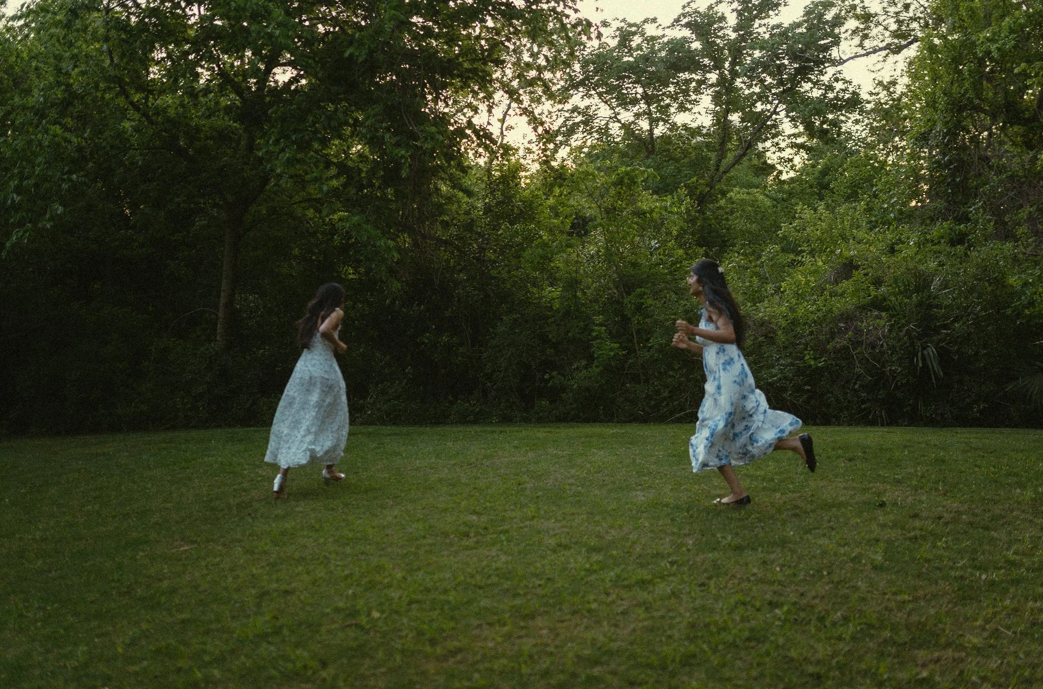 Two women in flowy dresses playing and running on a grassy area outdoors surrounded by trees during sunset.