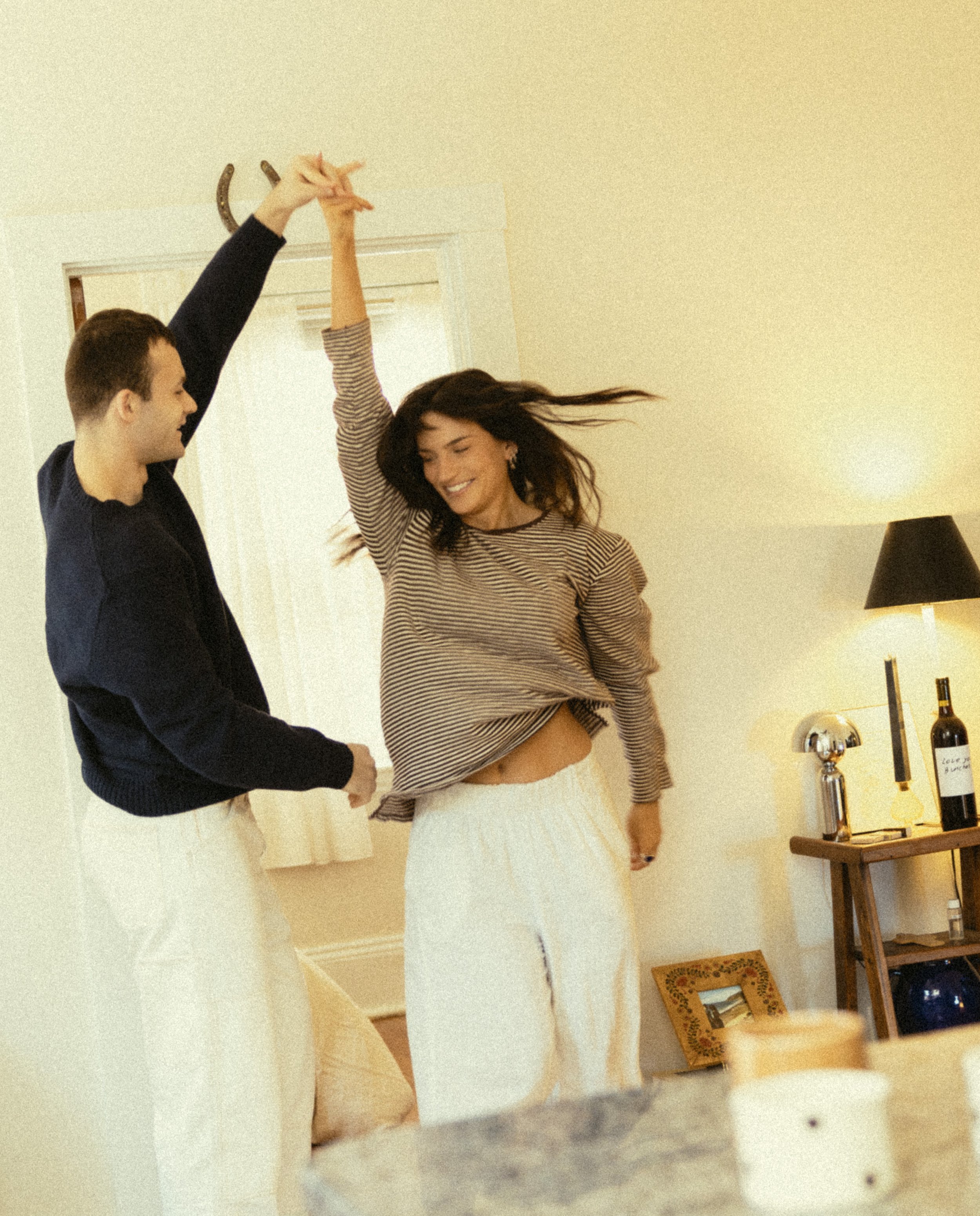 A woman with long dark hair wearing a striped sweater and a light-colored skirt dancing with a man with short dark hair wearing a dark sweater in a cozy, well-lit room with beige walls. A table with wine bottles and a lamp is visible in the backgroun