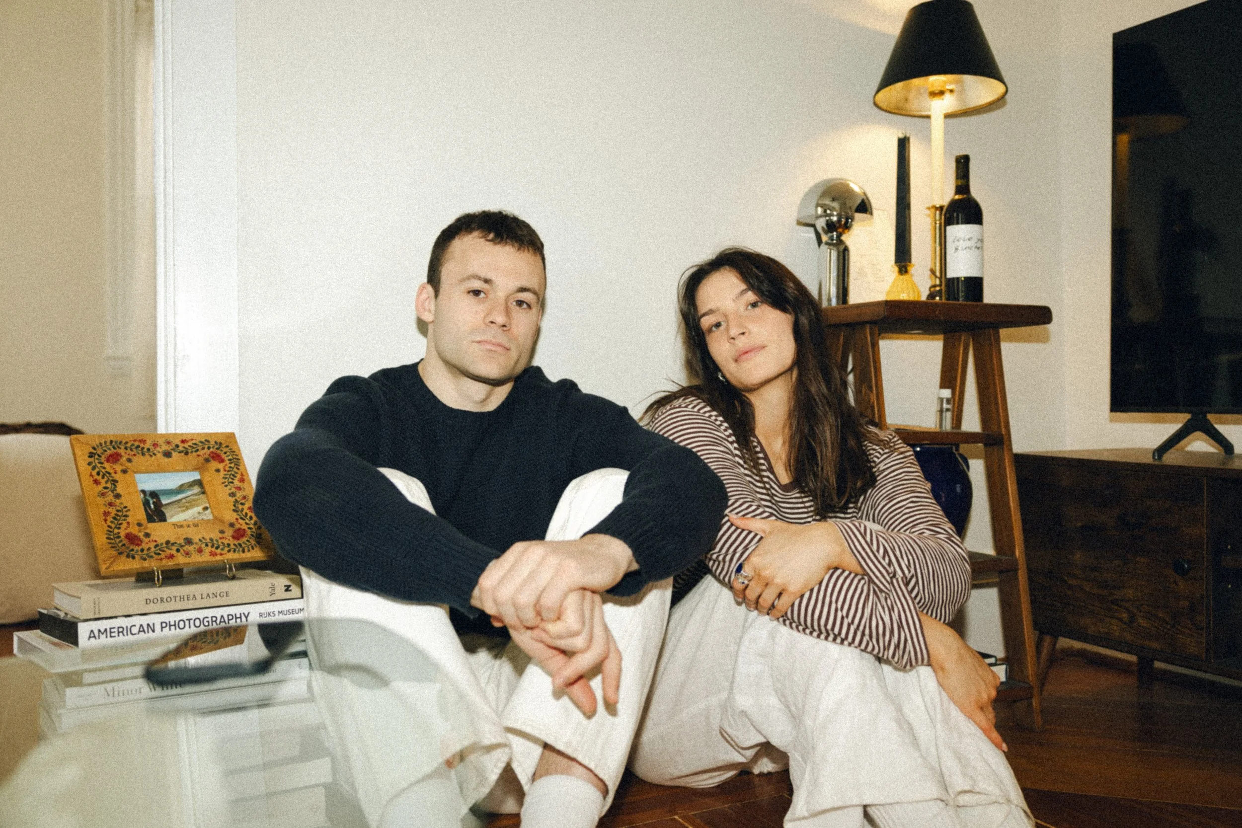 Two young adults, a man and a woman, sit closely together on a beige sofa in a living room. The man is wearing a black sweater and has short dark hair, while the woman is dressed in a striped shirt with long dark hair. Behind them is a wooden stand with a black lamp, a large bottle of wine, and a brass object. There are decorative books stacked on a side table, and a colorful picture frame with a floral border on the table.