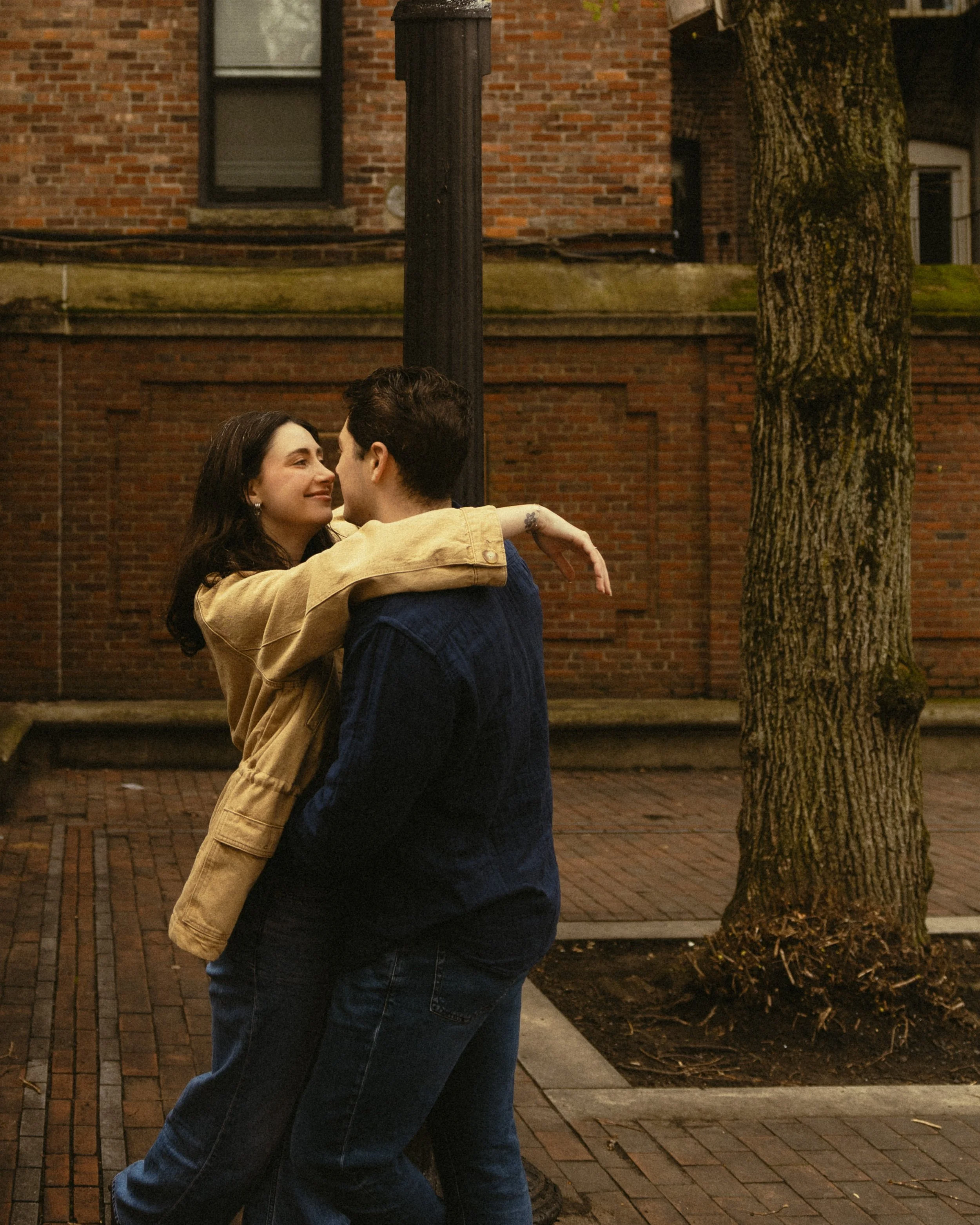 A young couple embracing each other and smiling at night in an urban setting, with a brick building, a leafless tree, and a streetlamp visible in the background.