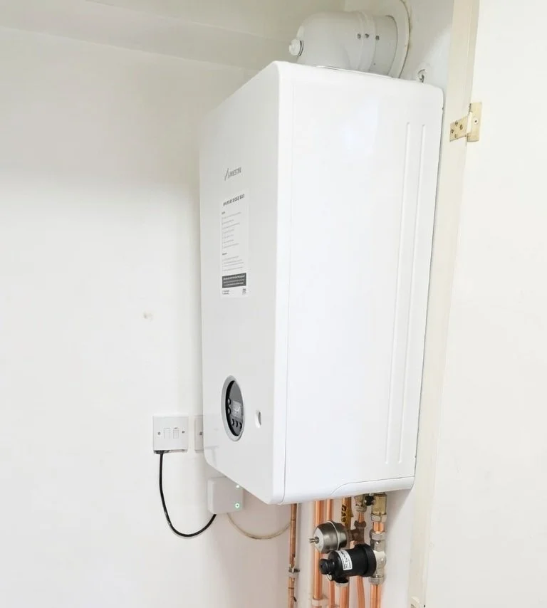 A wall-mounted white boiler with black control panel, copper pipes, and a vent pipe on a white wall.