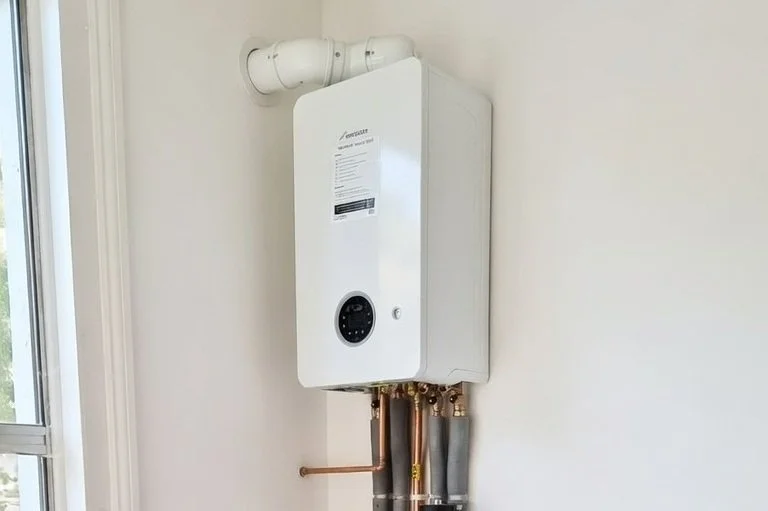 A wall-mounted tankless water heater with copper and gray pipes connected underneath, installed near a window in a white room.