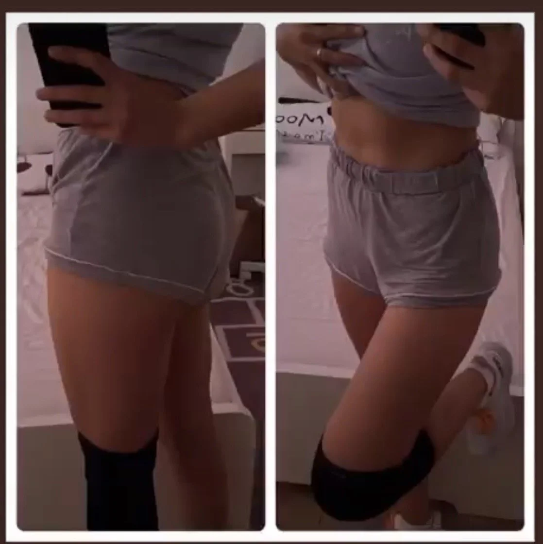 Side view of a woman in a gray t-shirt and gray shorts, taking mirror selfies to show her body transformation, with one knee on the bed, in a bedroom