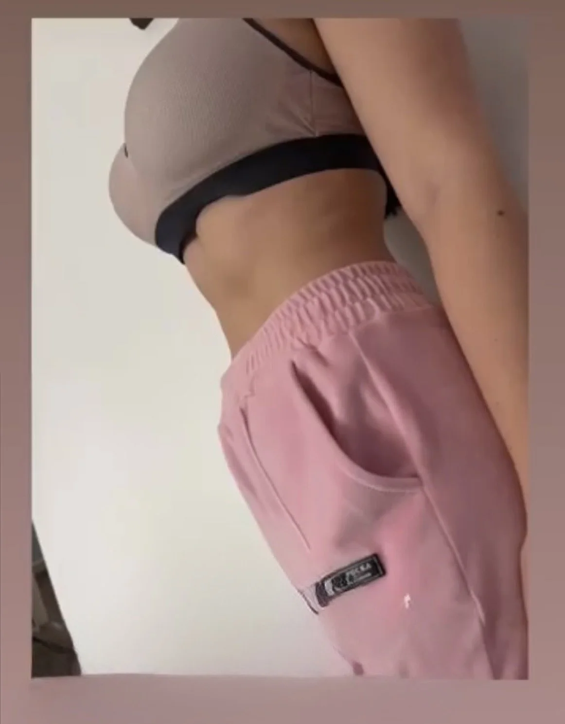 Close-up of a person's waist and side, wearing pink sweatpants and a cropped beige top, against a plain background.