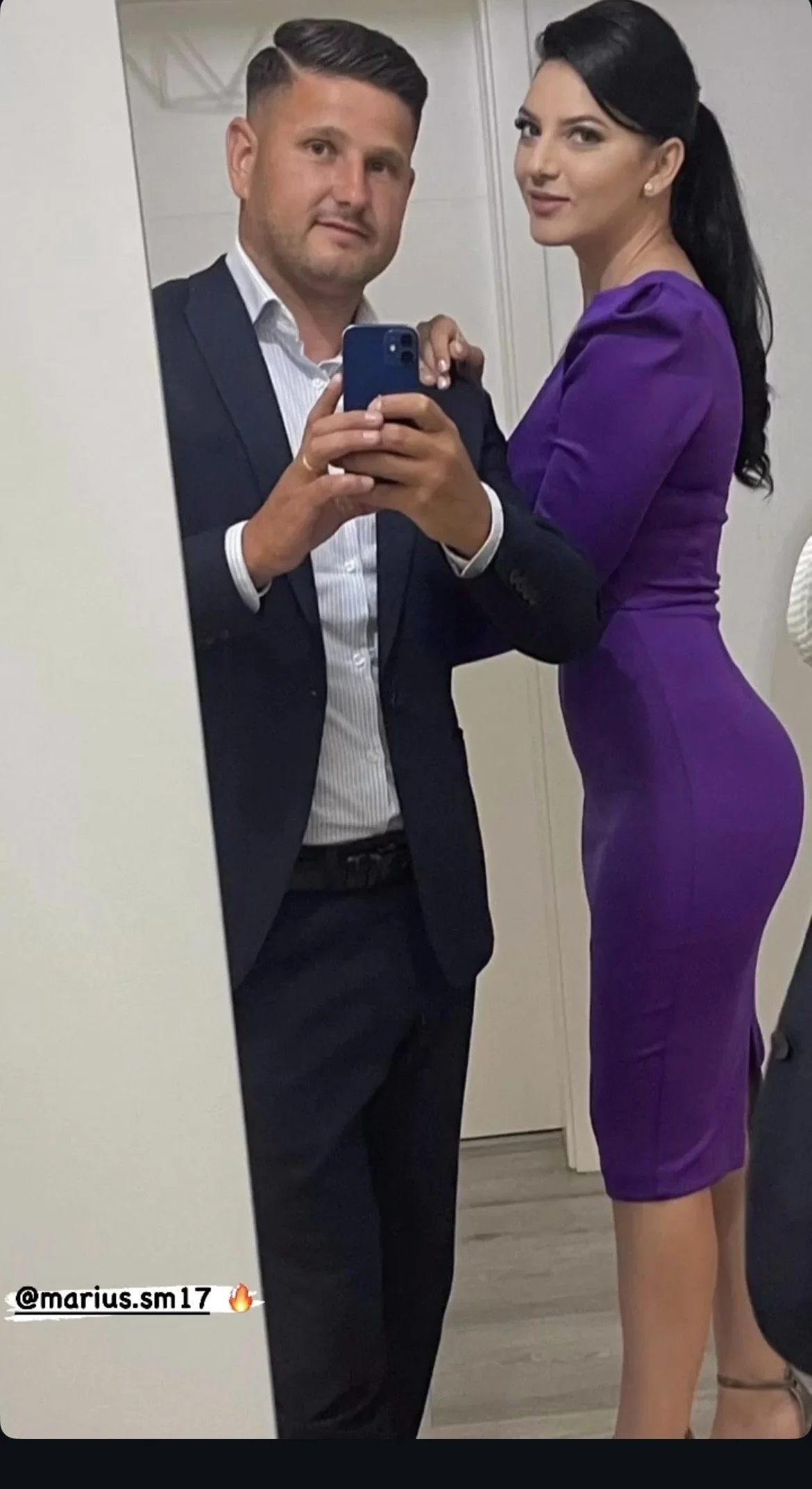 A man and woman take a mirror selfie in formal attire. The man wears a dark suit and white shirt, and the woman wears a fitted purple dress. They are indoors, likely at an event or gathering.