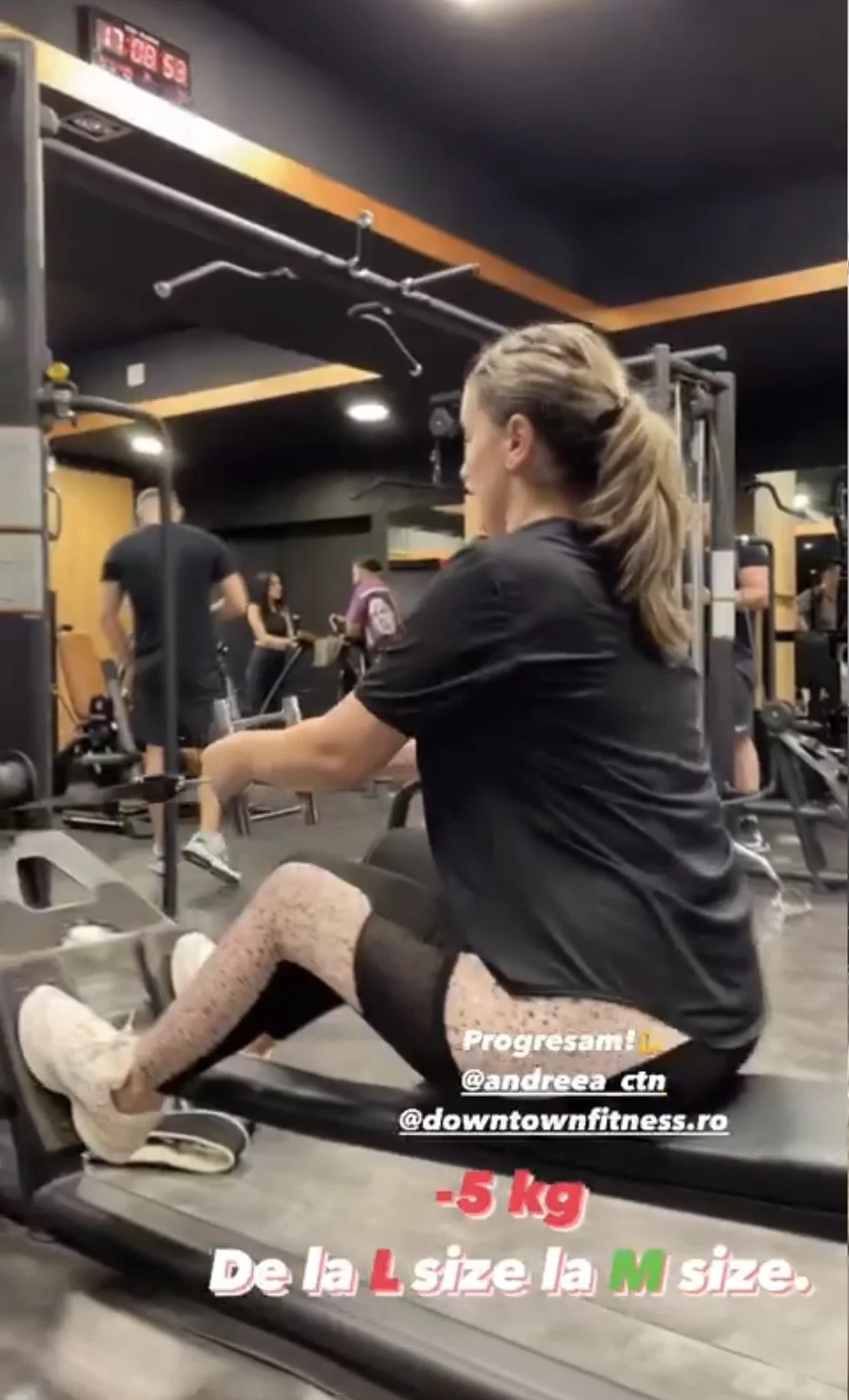 A woman with brown hair tied back in a ponytail, wearing a black t-shirt, patterned leggings, and sneakers, is sitting on a rowing machine in a gym. Other people are working out and walking around in the background. The gym has black walls with yello