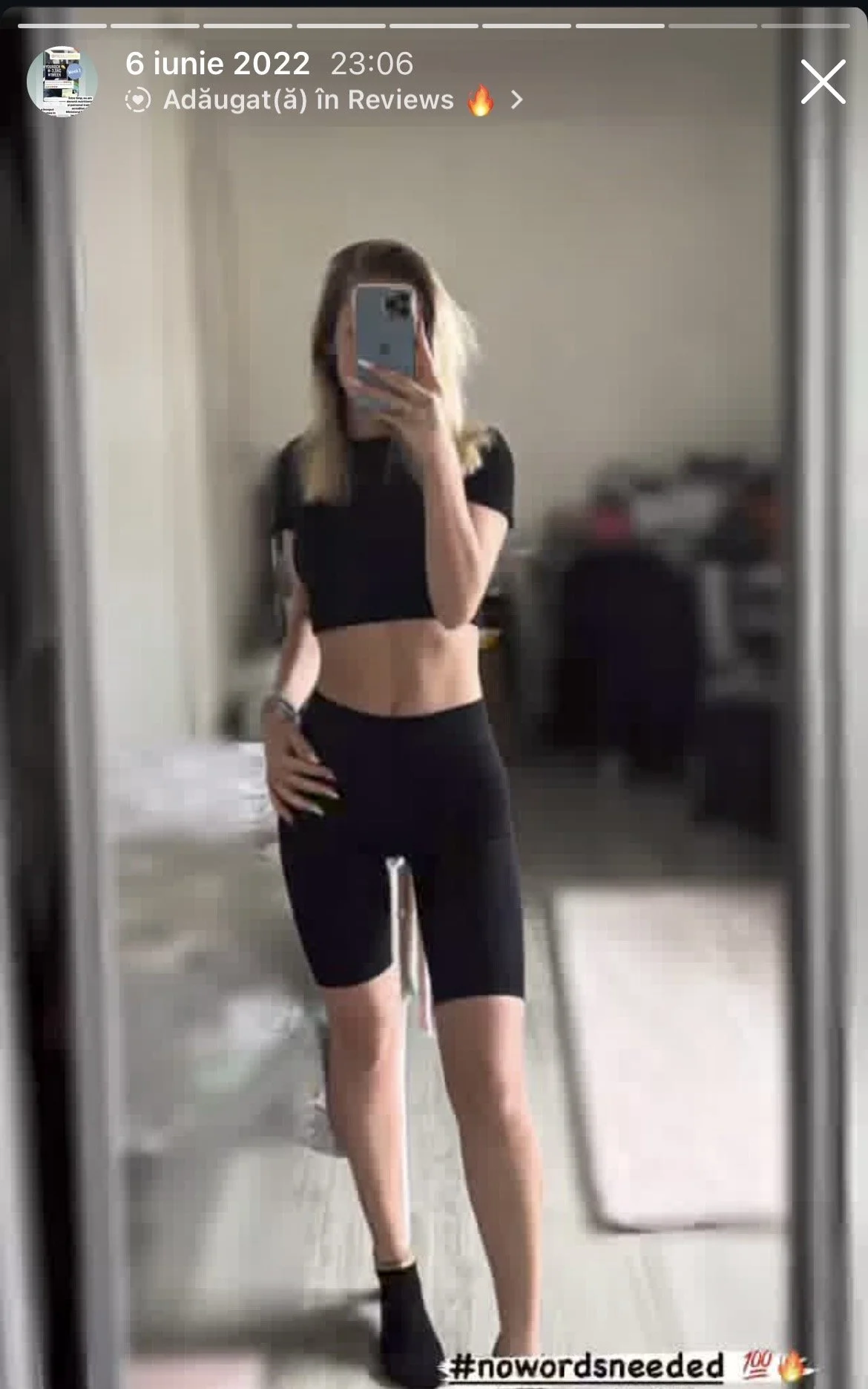 A woman taking a mirror selfie in a bedroom, wearing a black crop top and black shorts, with blurred background showing a bed, desk, and chair.