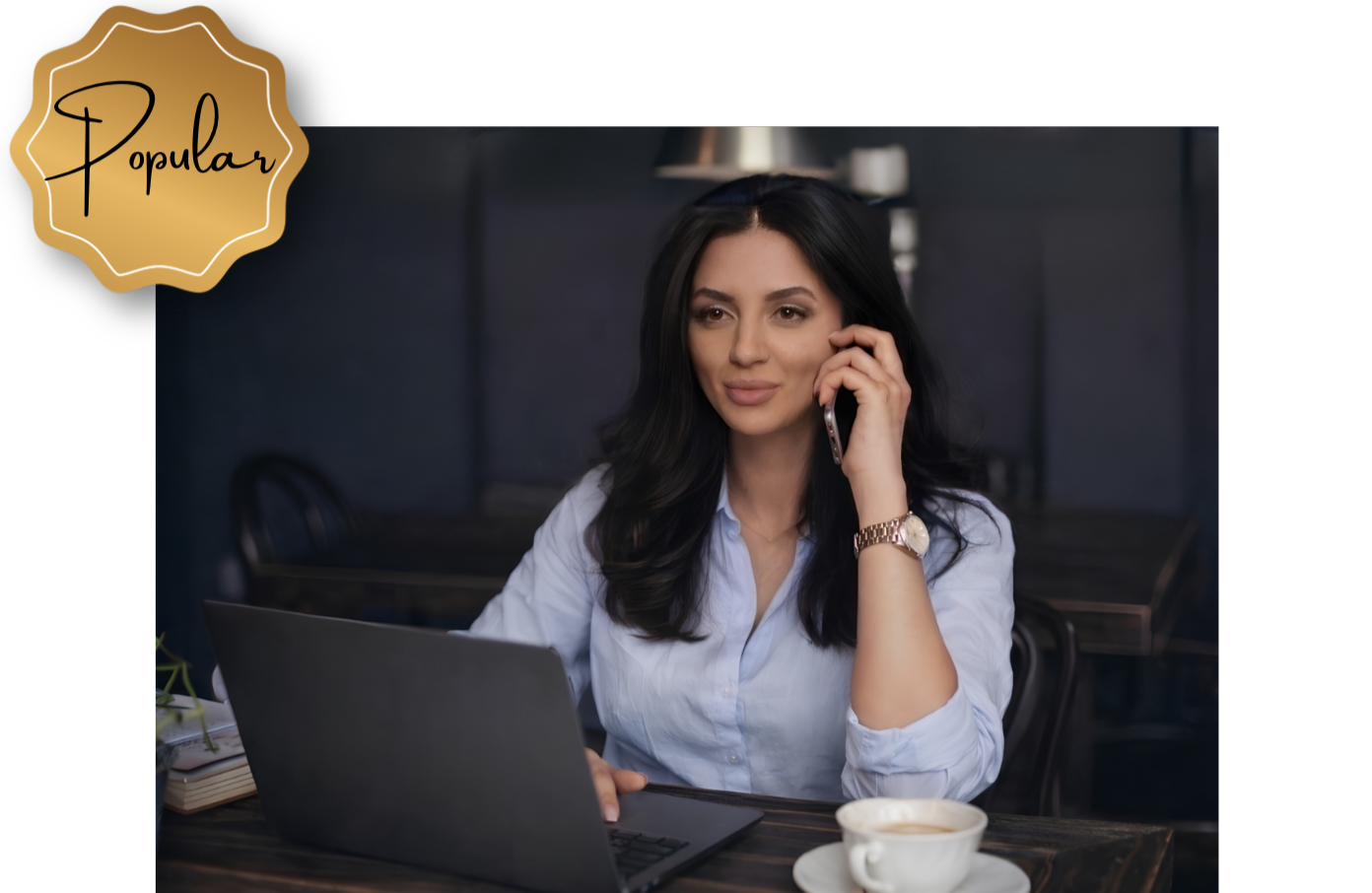 A woman with long dark hair, wearing a white shirt, is sitting at a dark wooden table in a cafe or office, talking on her mobile phone, with a laptop open in front of her, a cup of coffee, and a notebook nearby.