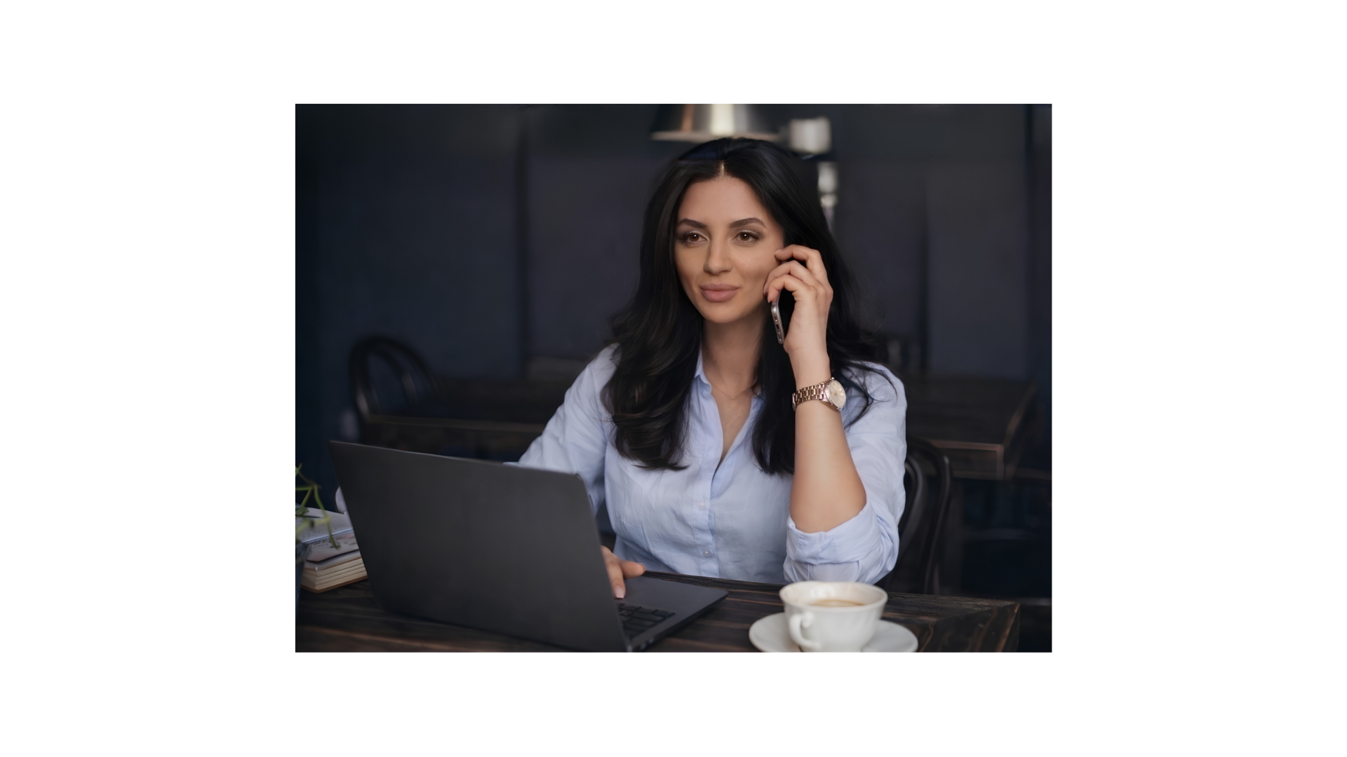 A woman with long black hair, wearing a white blouse and a gold watch, is sitting at a table with a laptop and a cup of coffee, talking on her cellphone.