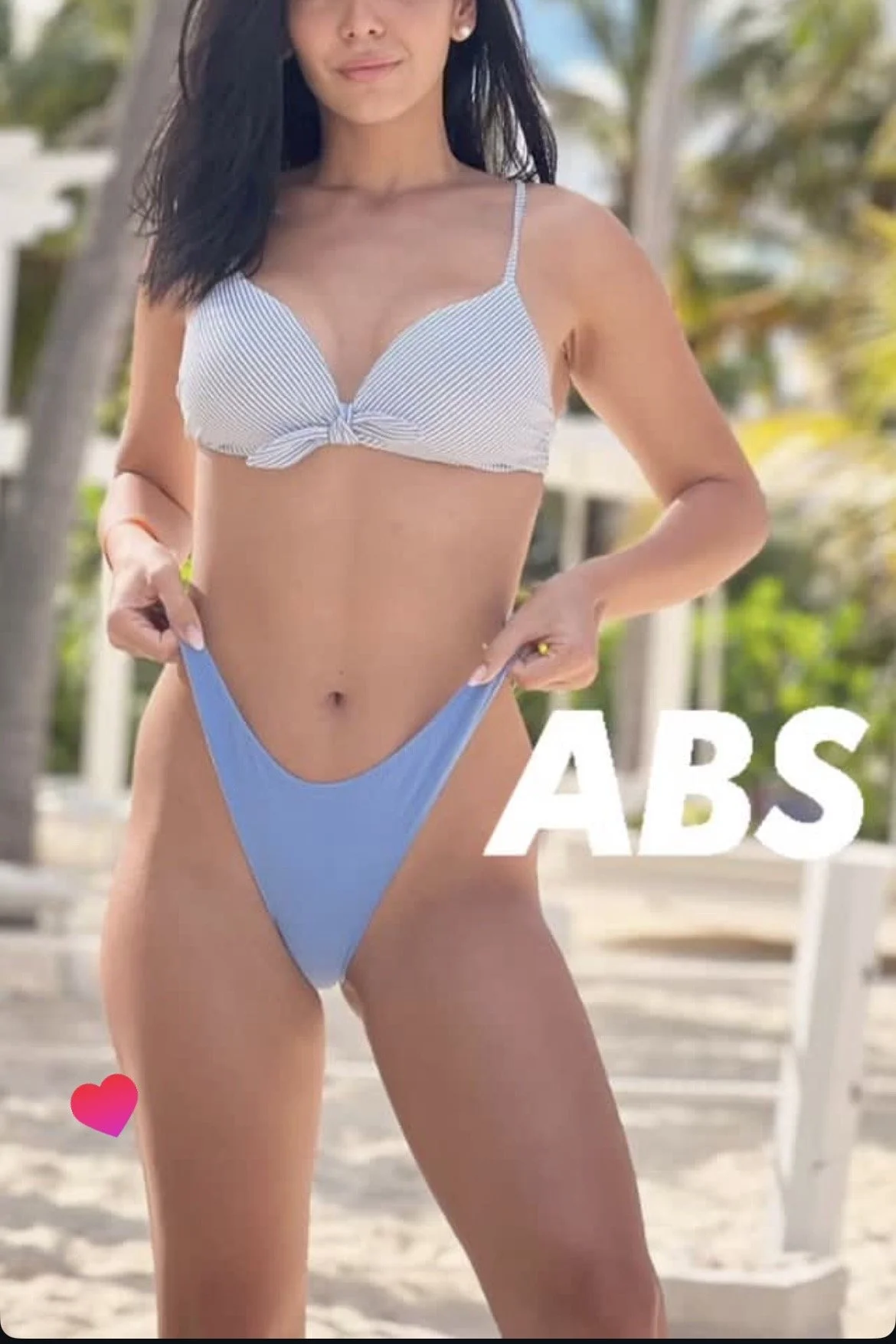 A woman standing on a beach with palm trees in the background, wearing a striped bikini top and blue bikini bottom, holding her bikini bottom with her hands, with the word 'ABS' written in large text.