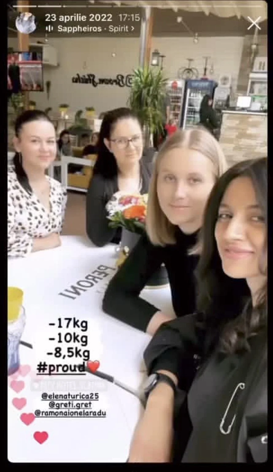 Four women sitting at a table in a restaurant, smiling at the camera. The table has a sign with weight loss progress: -17kg, -10kg, -8.5kg, and a hashtag #proud with a red heart emoji. One woman holds a bouquet of flowers, and there are potted plants