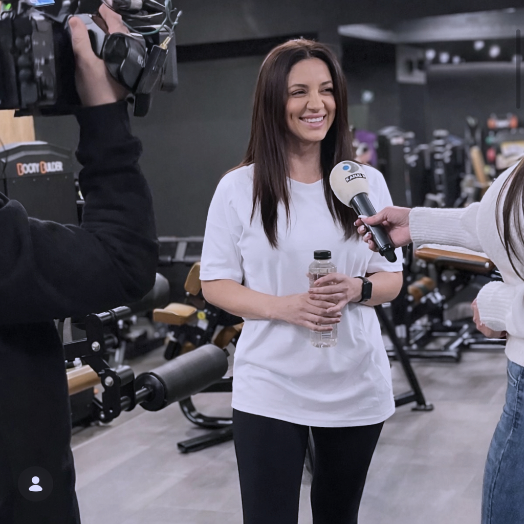 A woman being interviewed in a gym, holding a water bottle, smiling, with a camera operator filming her and a person holding a microphone with a news logo.