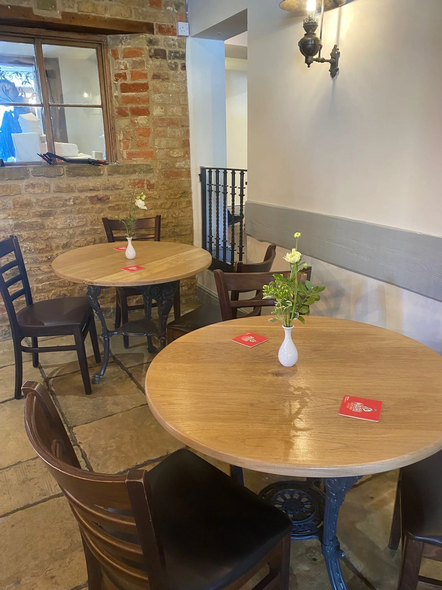Two round wooden tables with small vases holding single flowers, surrounded by wooden chairs in a cozy cafe or restaurant setting with exposed brick wall and white wall with a decorative light fixture.