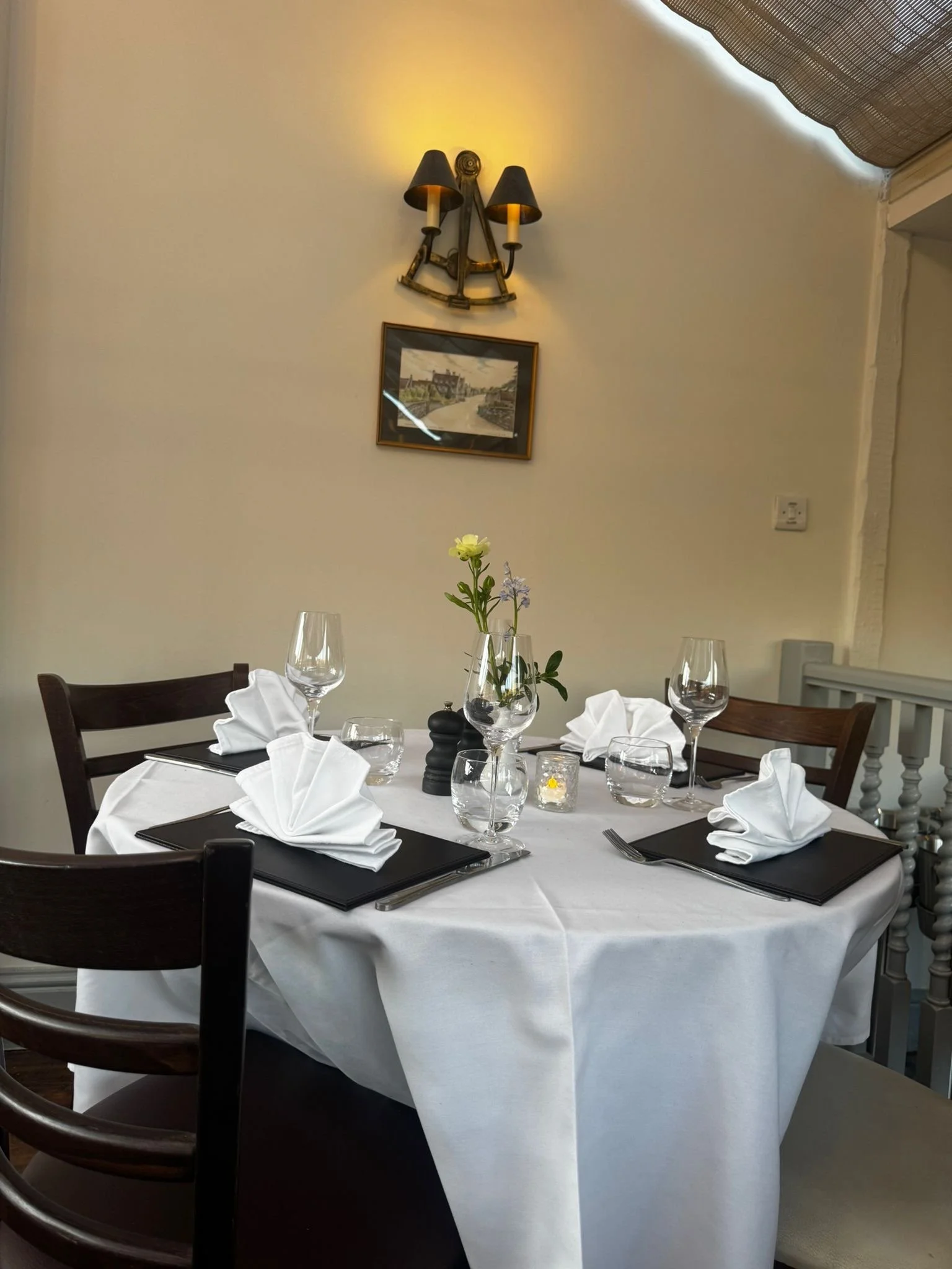 Set dining table with white tablecloth, black placemats, folded white napkins, wine glasses, water glasses, small candles, and a floral centerpiece in a restaurant or dining area.