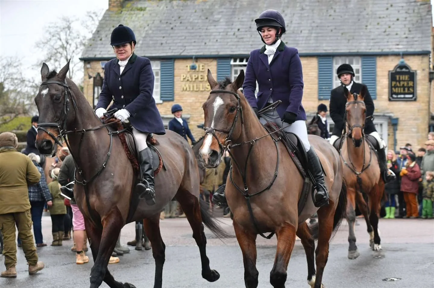 People riding horses in a parade in front of a crowd, with a building labeled 'The Paper Mills' in the background.