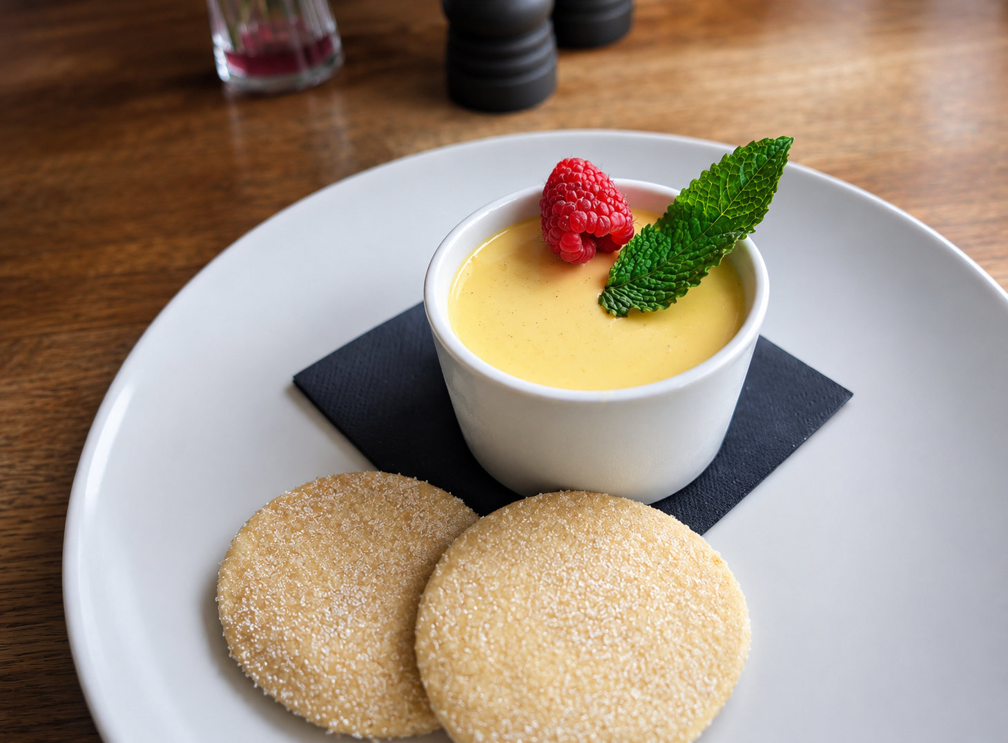 Creamy custard dessert garnished with a raspberry and a mint leaf, served with two sugar-dusted cookies on a white plate.