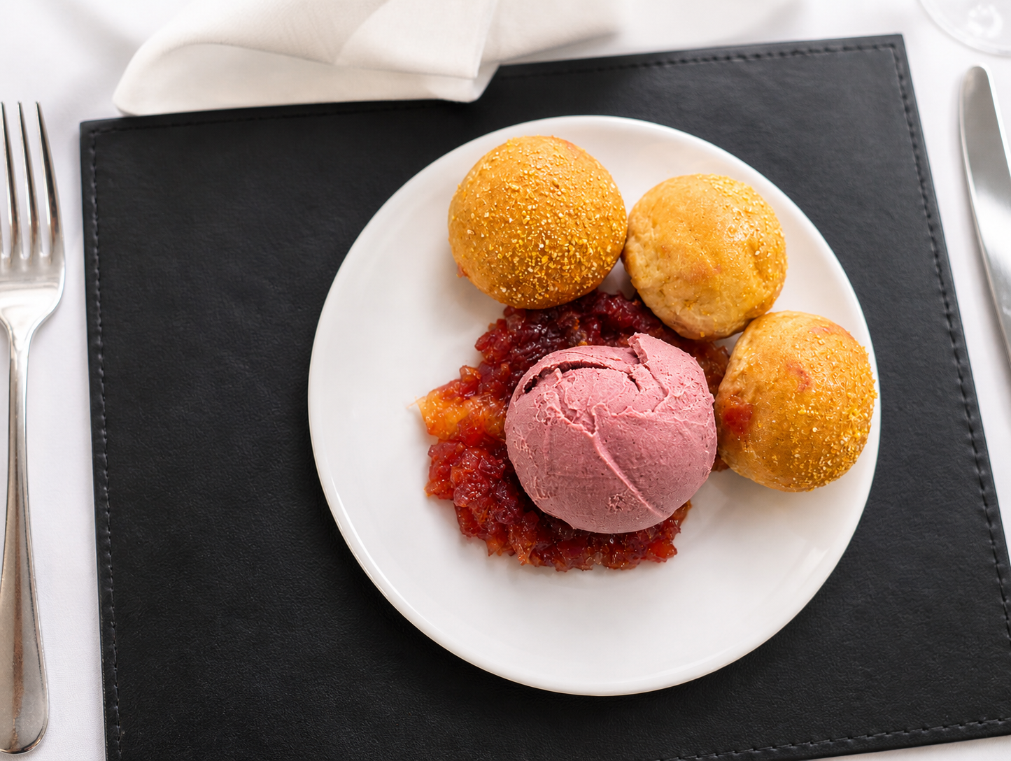 Plate of three fried cheese balls, a scoop of pink ice cream or sorbet, and a serving of fruit compote, on a black placemat with a fork and knife nearby.
