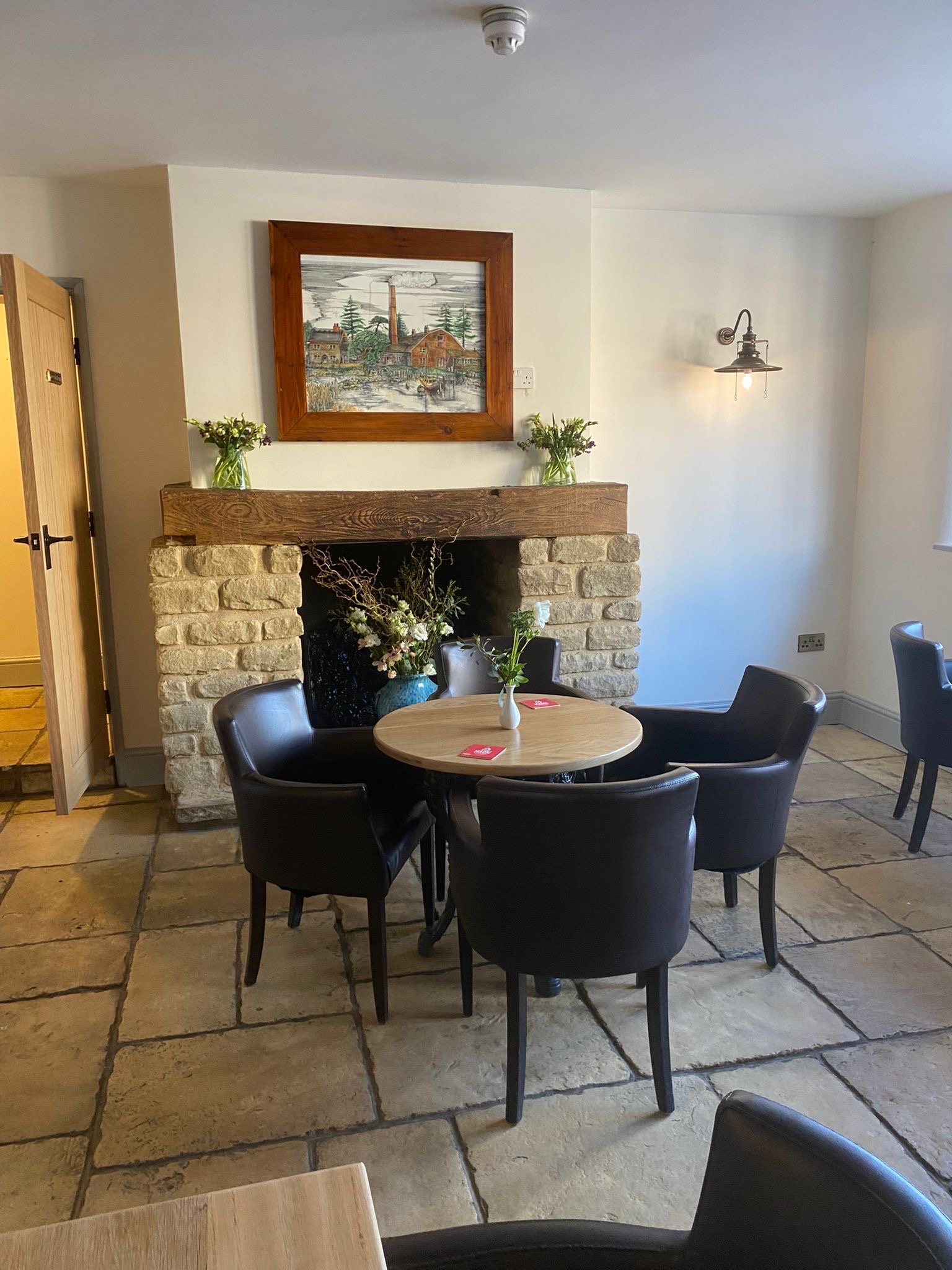A cozy dining area with a small round wooden table surrounded by five black leather chairs, a decorated stone fireplace with flowers and a framed painting above, and a window with a wall-mounted light on the right.