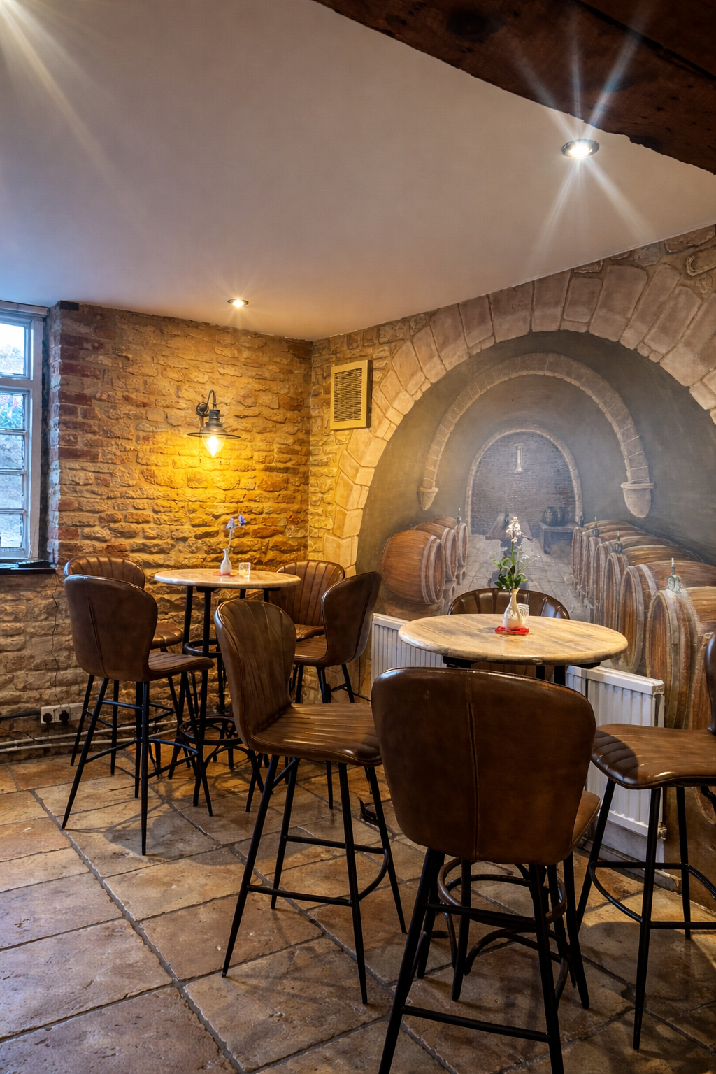 Interior of a cozy restaurant with brick walls, wooden tables with vases, leather chairs, a mural of a wine cellar, and warm lighting.