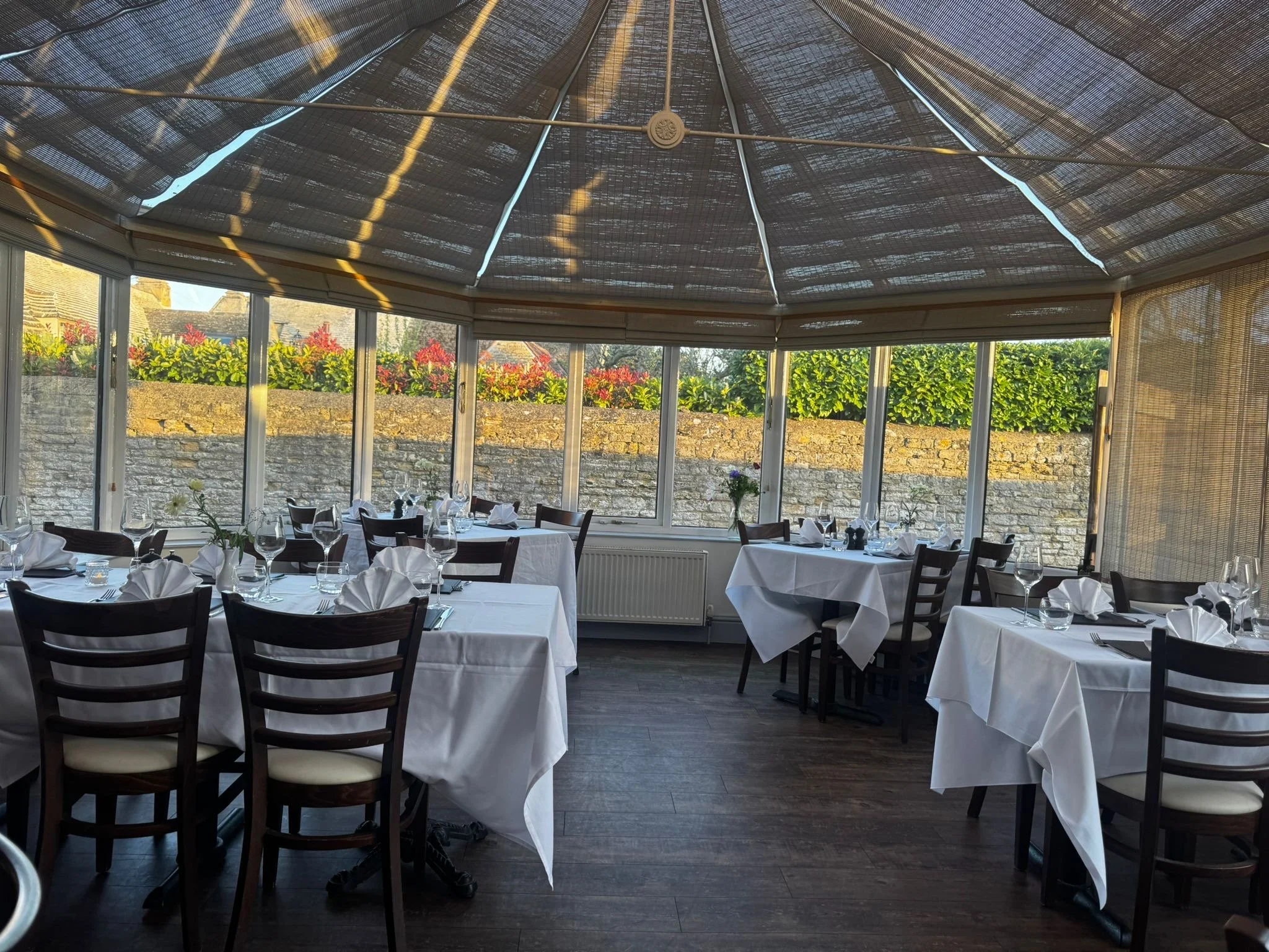 A restaurant with white tablecloths and dark wood chairs, set with wine glasses, napkins, and small vases, inside a glass-enclosed space with a view of a stone wall and garden outside.