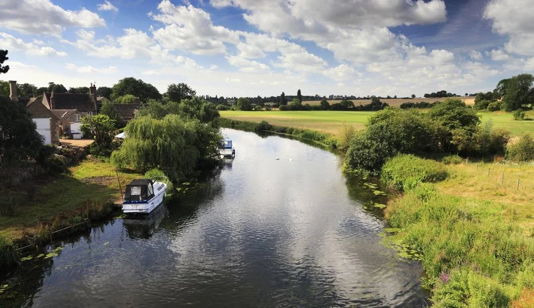 A scenic view of a river with boats, surrounded by lush greenery and fields, under a partly cloudy sky.
