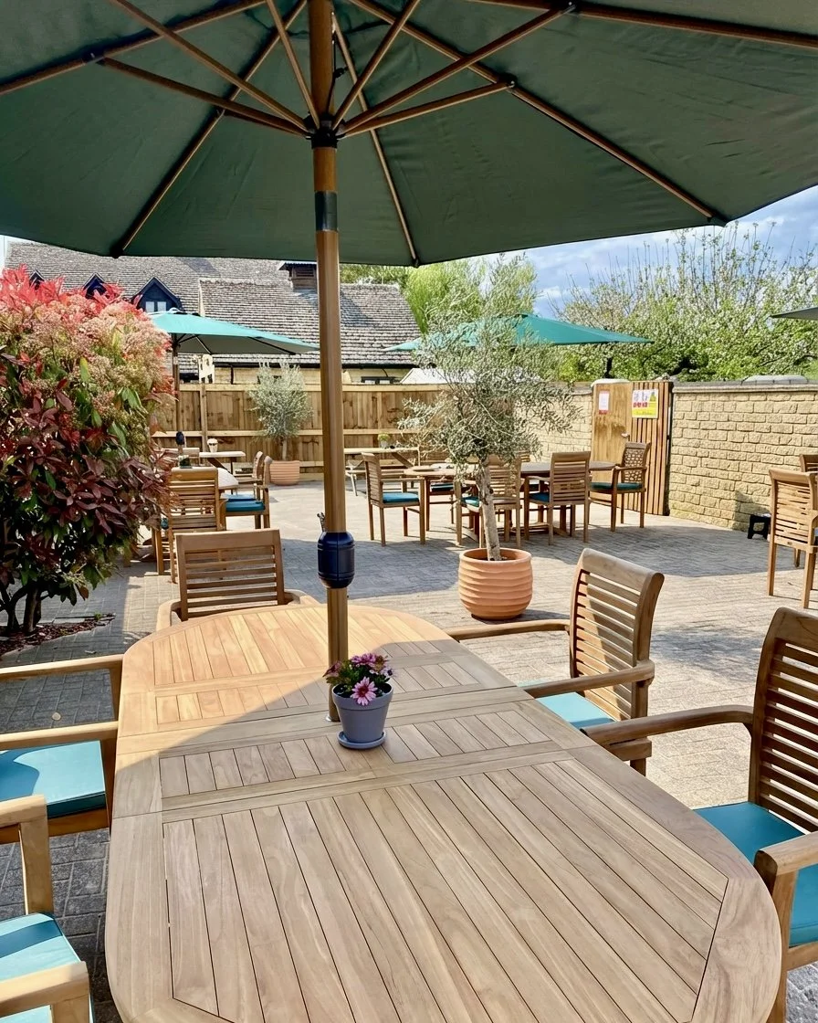 An outdoor patio area with wooden tables and chairs, large green patio umbrellas, potted plants, and a wooden fence, on a sunny day.