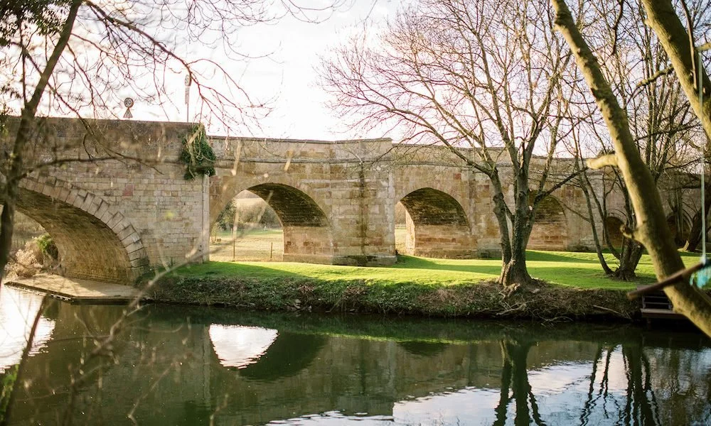 A historic stone bridge with multiple arches over a calm river, surrounded by leafless trees in late autumn or winter.