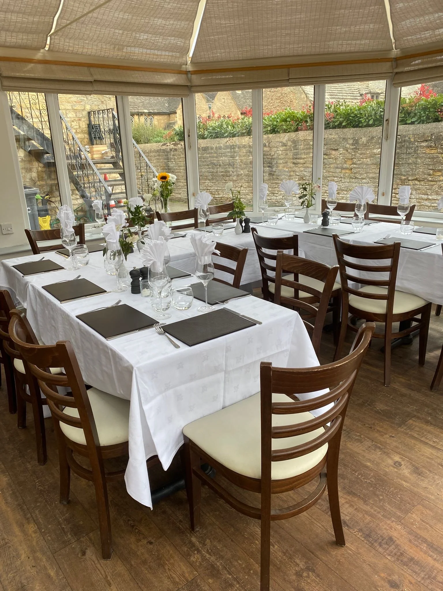 A restaurant or dining room with tables set for a meal, featuring white tablecloths, napkins, glasses, plates, and flower arrangements, including a sunflower. Large windows provide a view of outdoors with stone walls and garden plants.