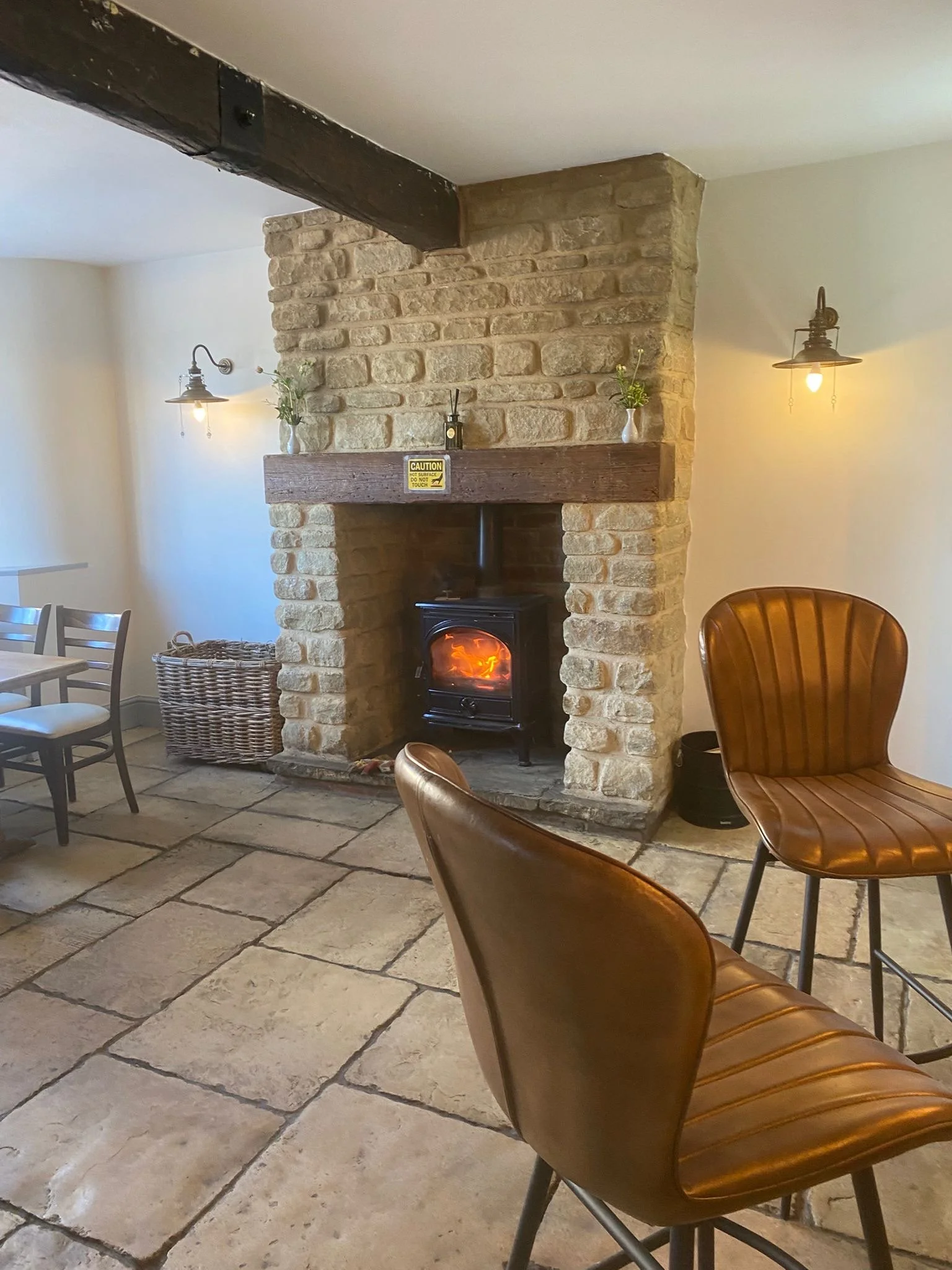 Interior of a cozy room with a stone fireplace, a wood stove, and a wooden beam. There are brown leather chairs, a small table, a basket, and wall-mounted lamps. The fireplace has a sign that says "CAUTION." The floor is made of large stone tiles.