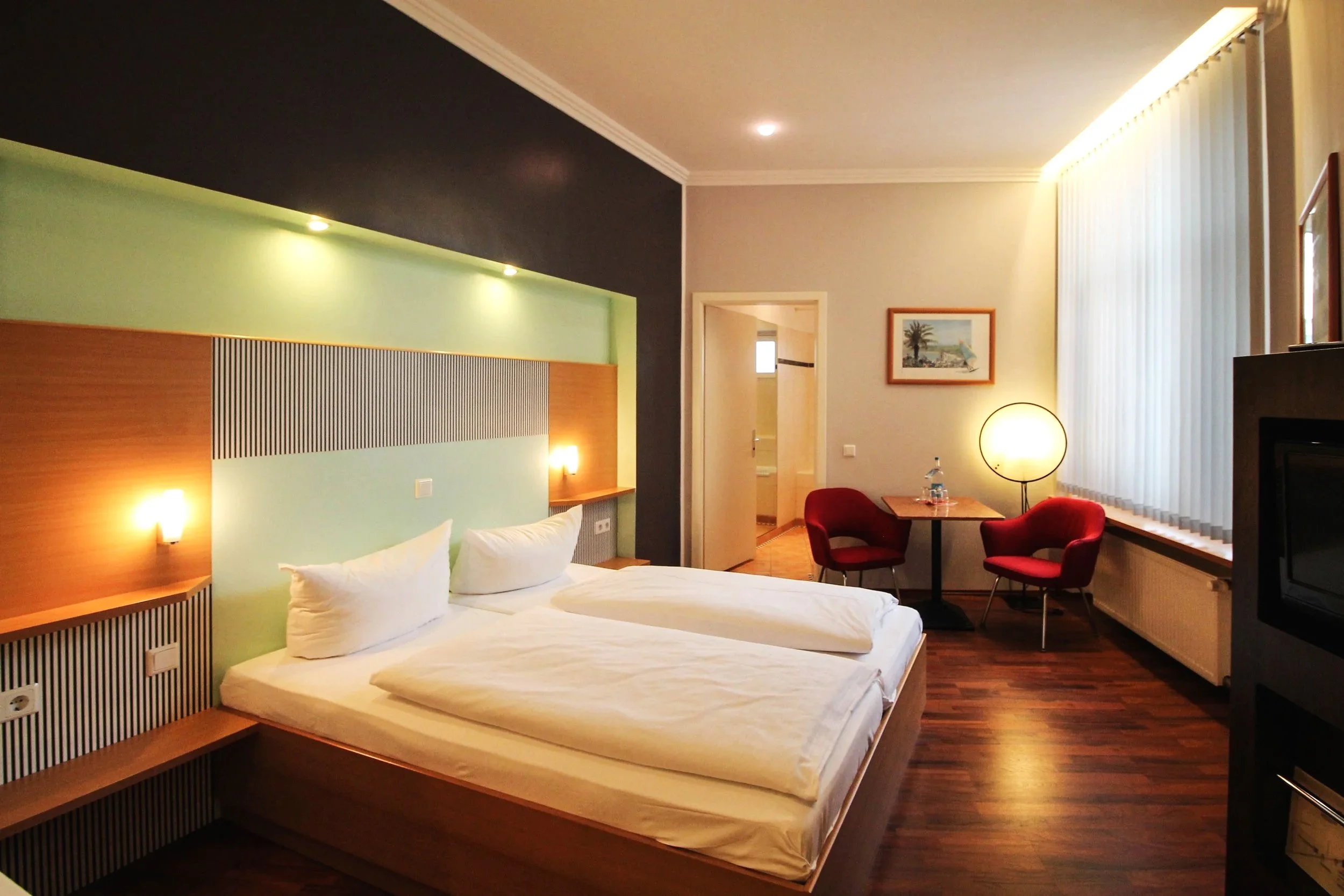 Ein Hotelzimmer mit einem Doppelbett, einem Sessel, einem Tisch, einem Lampenschirm und einem Gemälde an der Wand.
