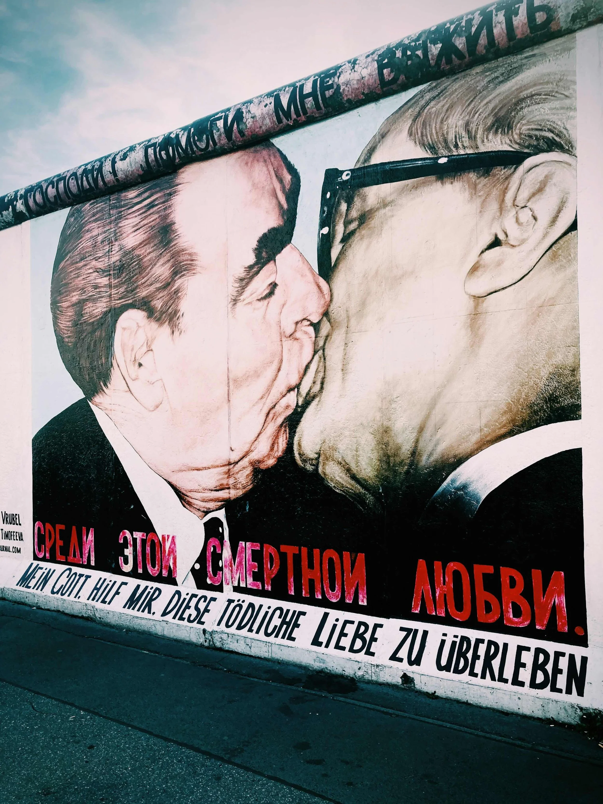 Straßenkunst mit einem Wandgemälde, das zwei Männer zeigt, die sich küssen, mit Text in mehreren Sprachen darunter.