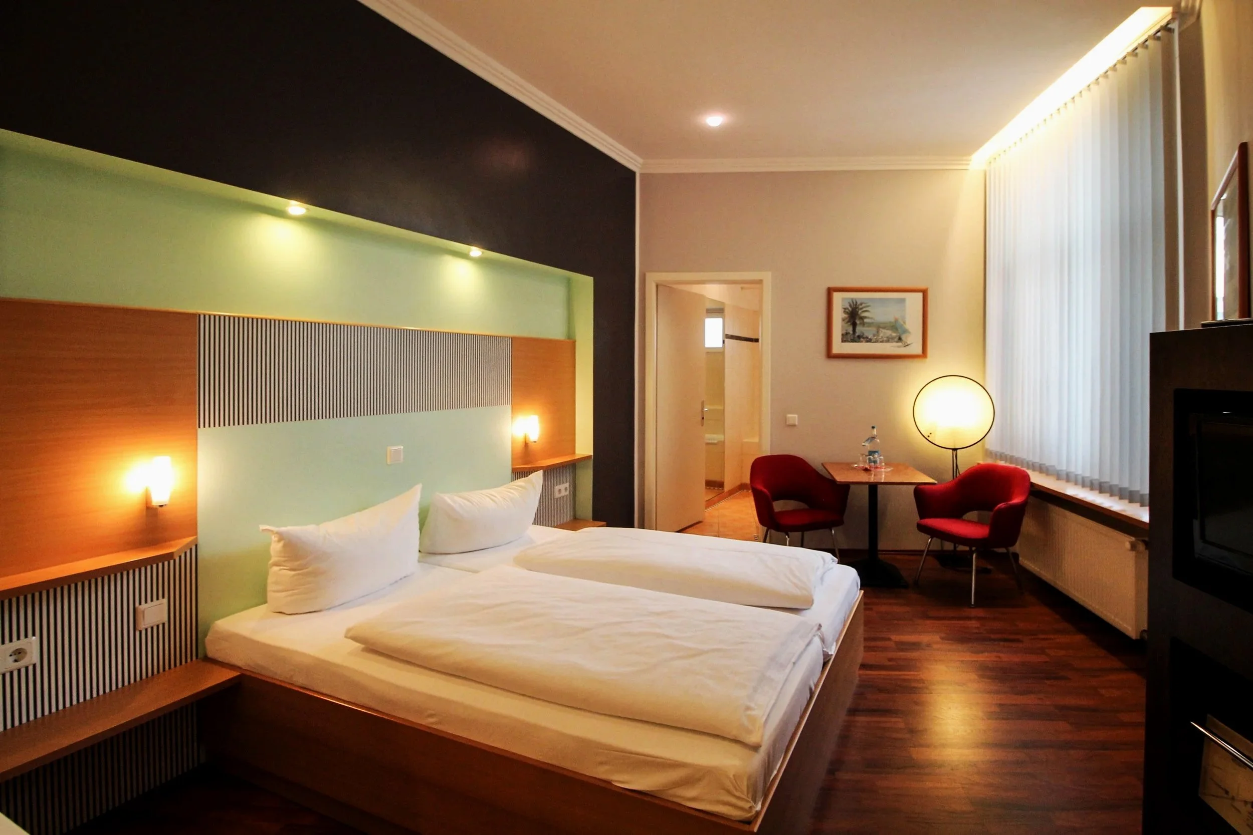 Ein Hotelzimmer mit Doppelbett, Sitzbereich mit zwei roten Stühlen, Schreibtisch, einer Lampe, und einem Fenster mit vertikalen Jalousien.