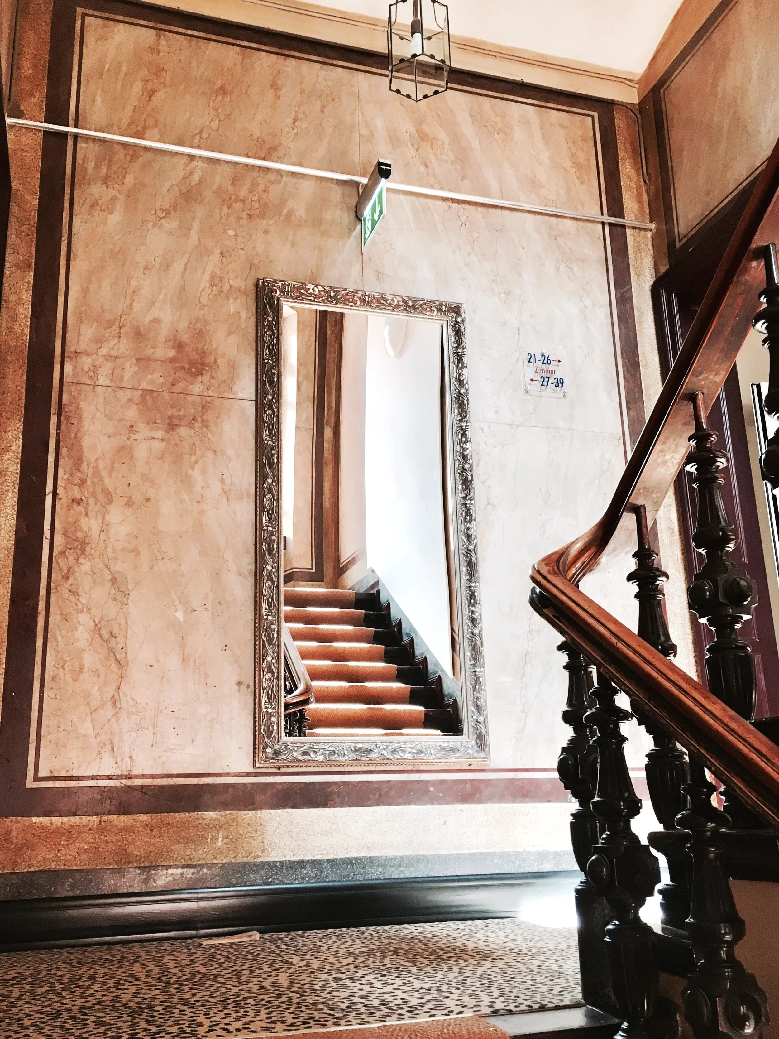 Ein Blick auf eine Treppe mitbraunen Stufen, einem großen rechteckigen Spiegel mit verzierter silberner Rahmen und einem Wandbild, das die Zimmertypen 21-26 und 27-39 in einem Gebäude anzeigt. Das Geländer ist aus dunklem Holz, und es gibt einen hoch