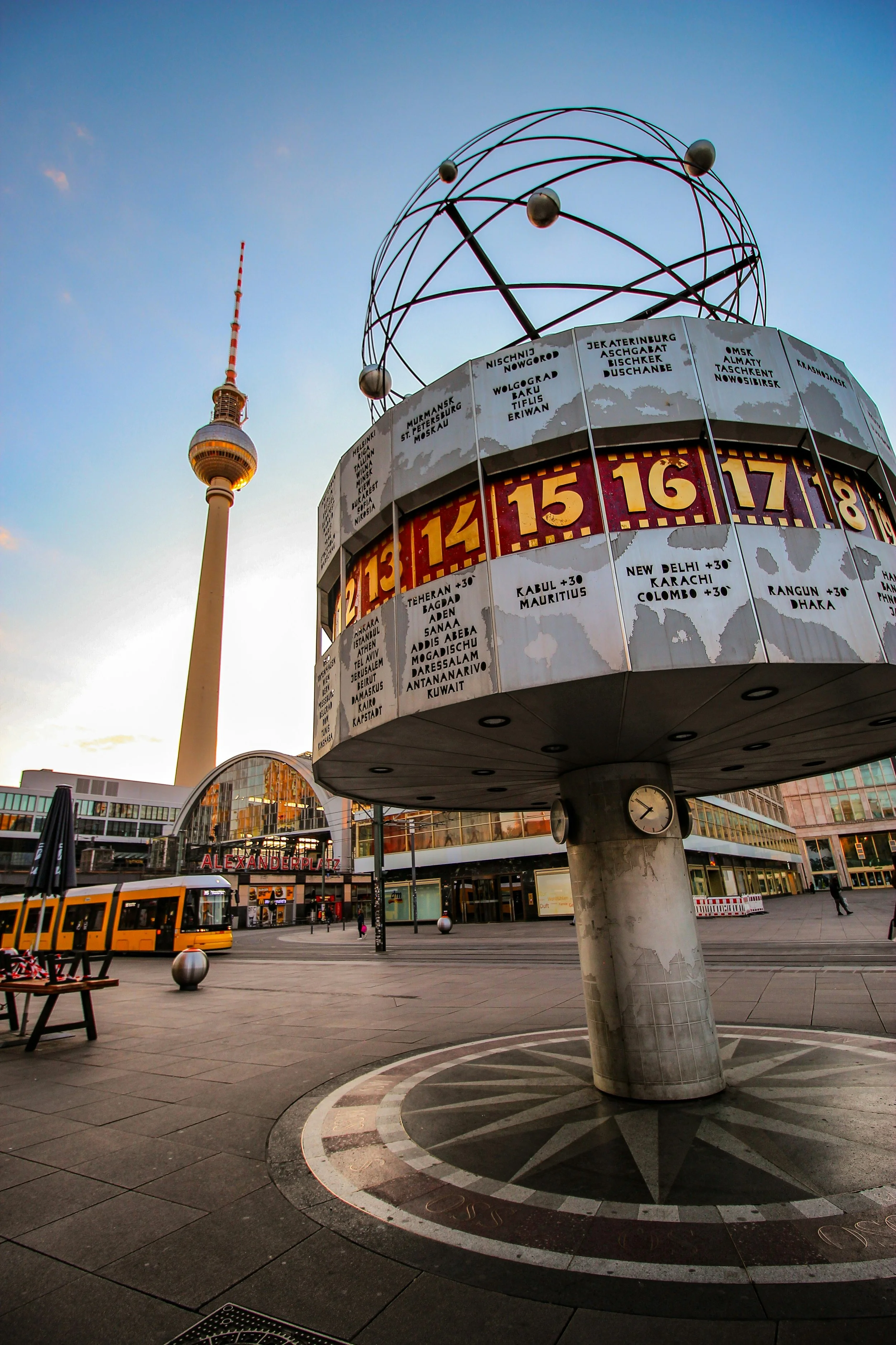 Der Berliner Alexanderplatz, mit einem großen Pylon, einem wettergeformten Globus aus Metall, einer U-Bahn mit gelben Waggons, einem Gebäude mit einer Glasfassade und einem Eingang mit dem Schriftzug 'ALEXANDERPLATZ'.