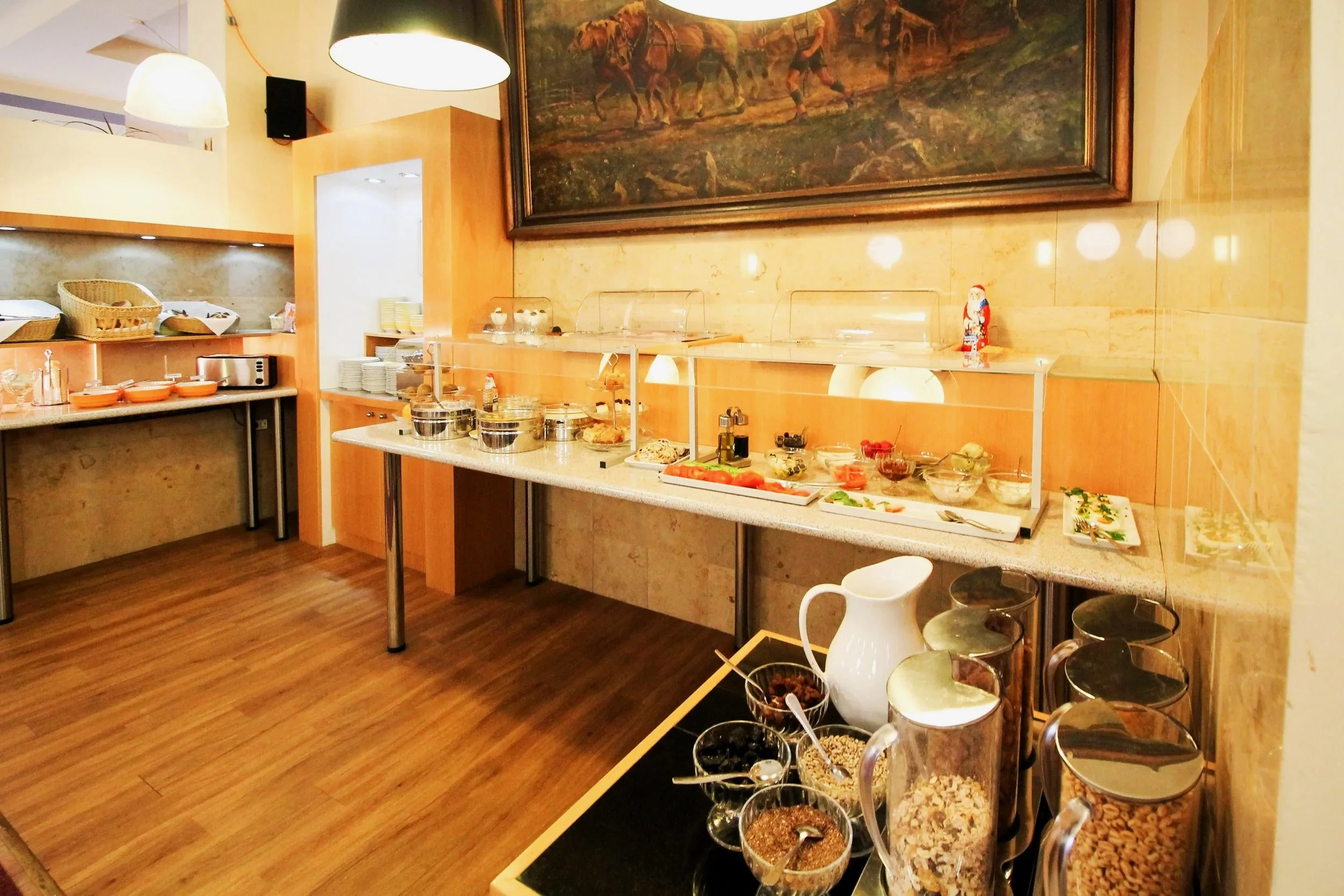 Frühstücksbuffet mit Aufschnitt, Marmeladen, Joghurt, Cerealien und Kuchen in einem Restaurant. Hängende Lampen, große Malerei an der Wand, Holztöne.