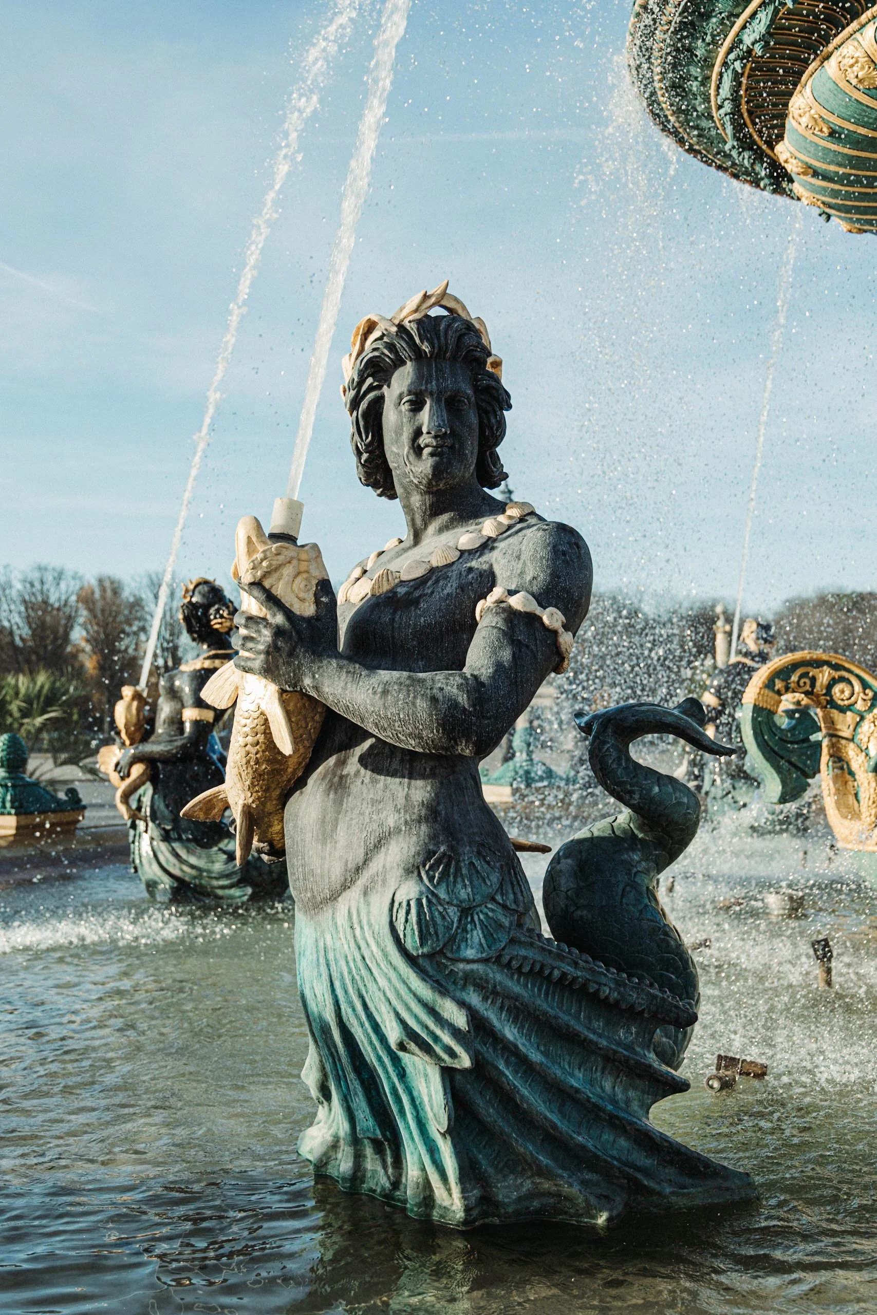 A statue of a mermaid holding a fish in a fountain with water splashing, surrounded by other maritime sculptures and water features.