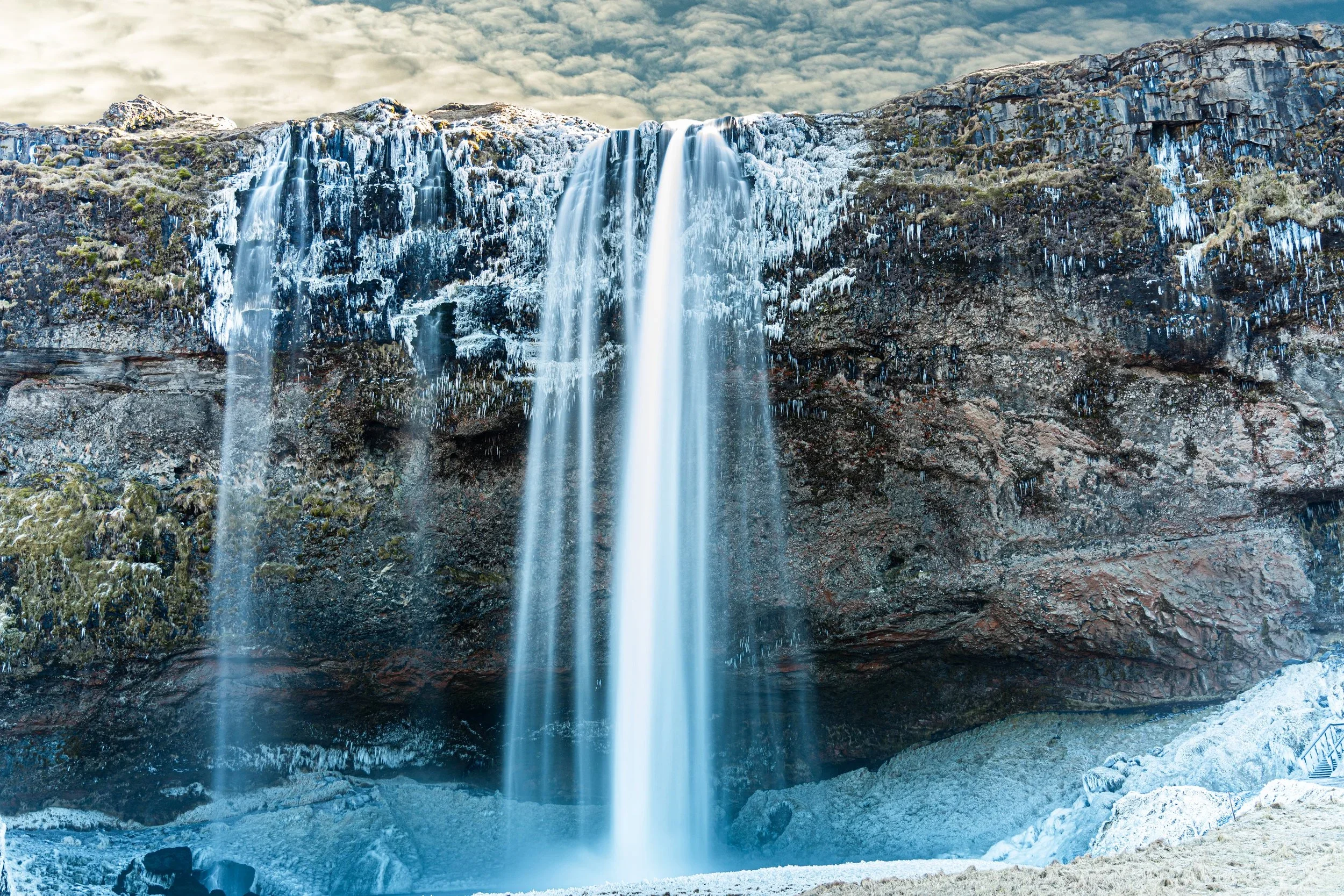 A waterfall flowing over a rocky cliff with patches of ice, icy terrain at the base, and a cloudy sky overhead.