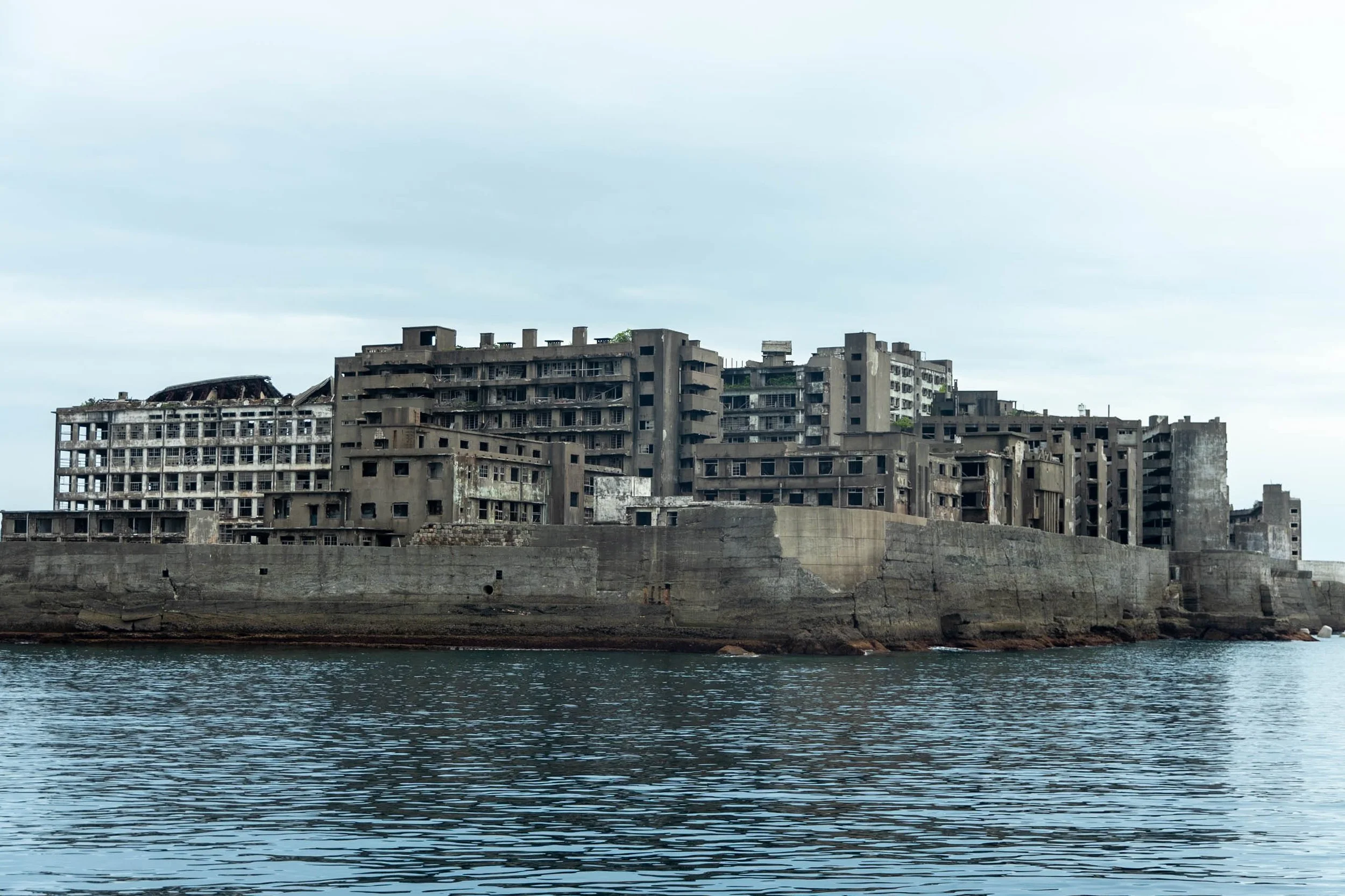 Abandoned, dilapidated concrete building on a waterfront, with broken windows and deteriorating structures under a cloudy sky.