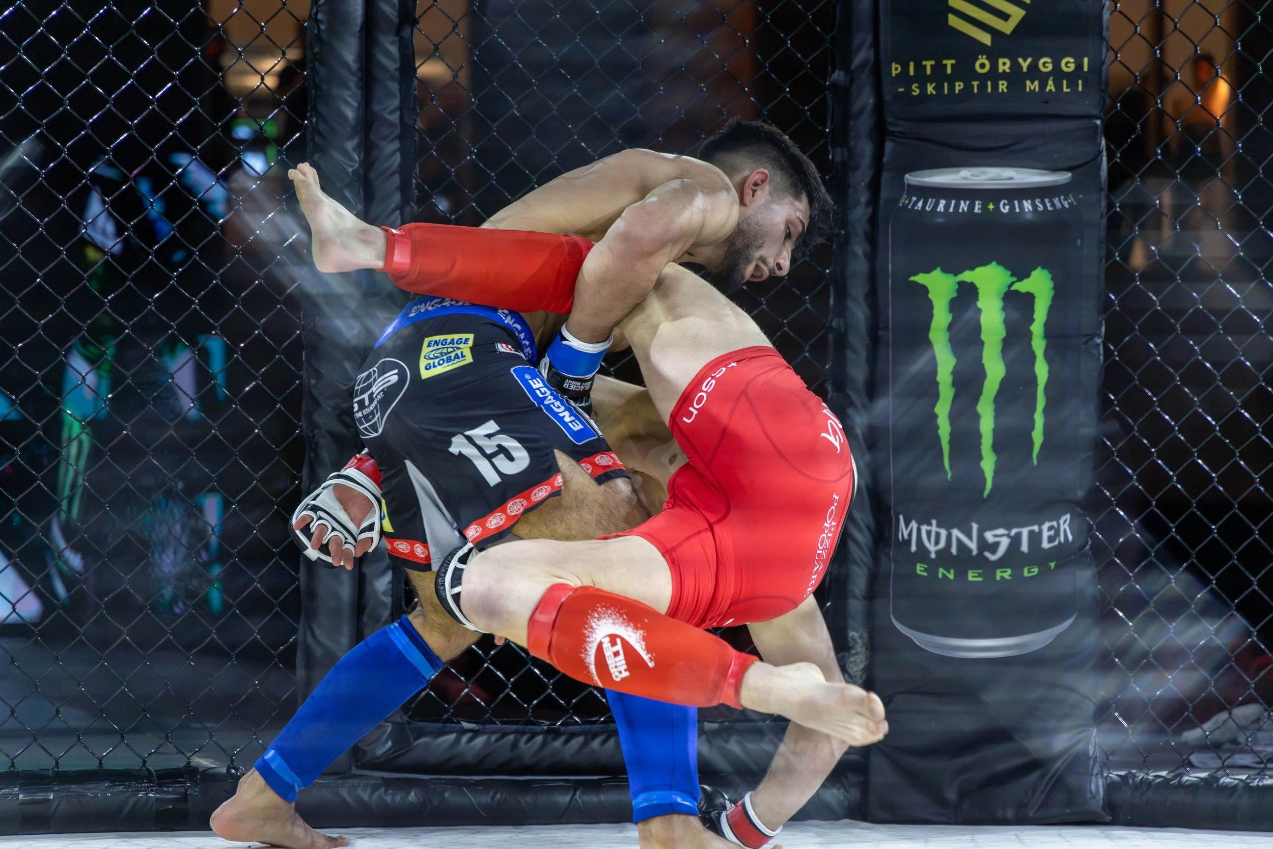 Two mixed martial artists fighting inside an octagon cage, with one fighter in red shorts and the other in black shorts, near a Monster Energy advertisement.