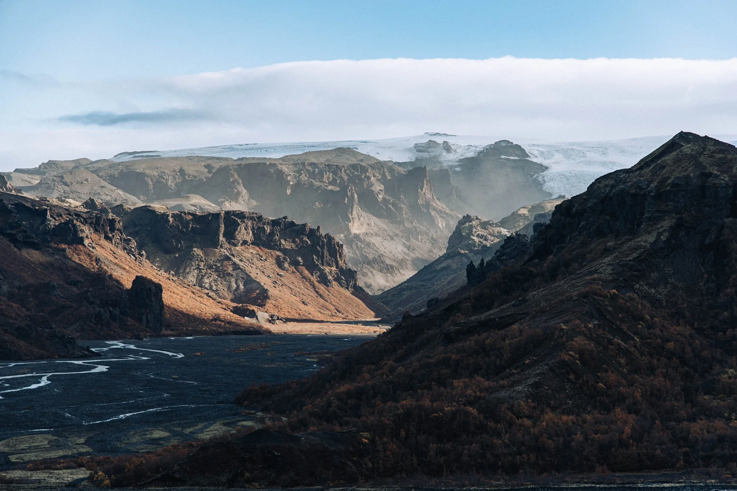 Mountain landscape with rugged cliffs, a valley, and a river, with snow-capped peaks in the distance.