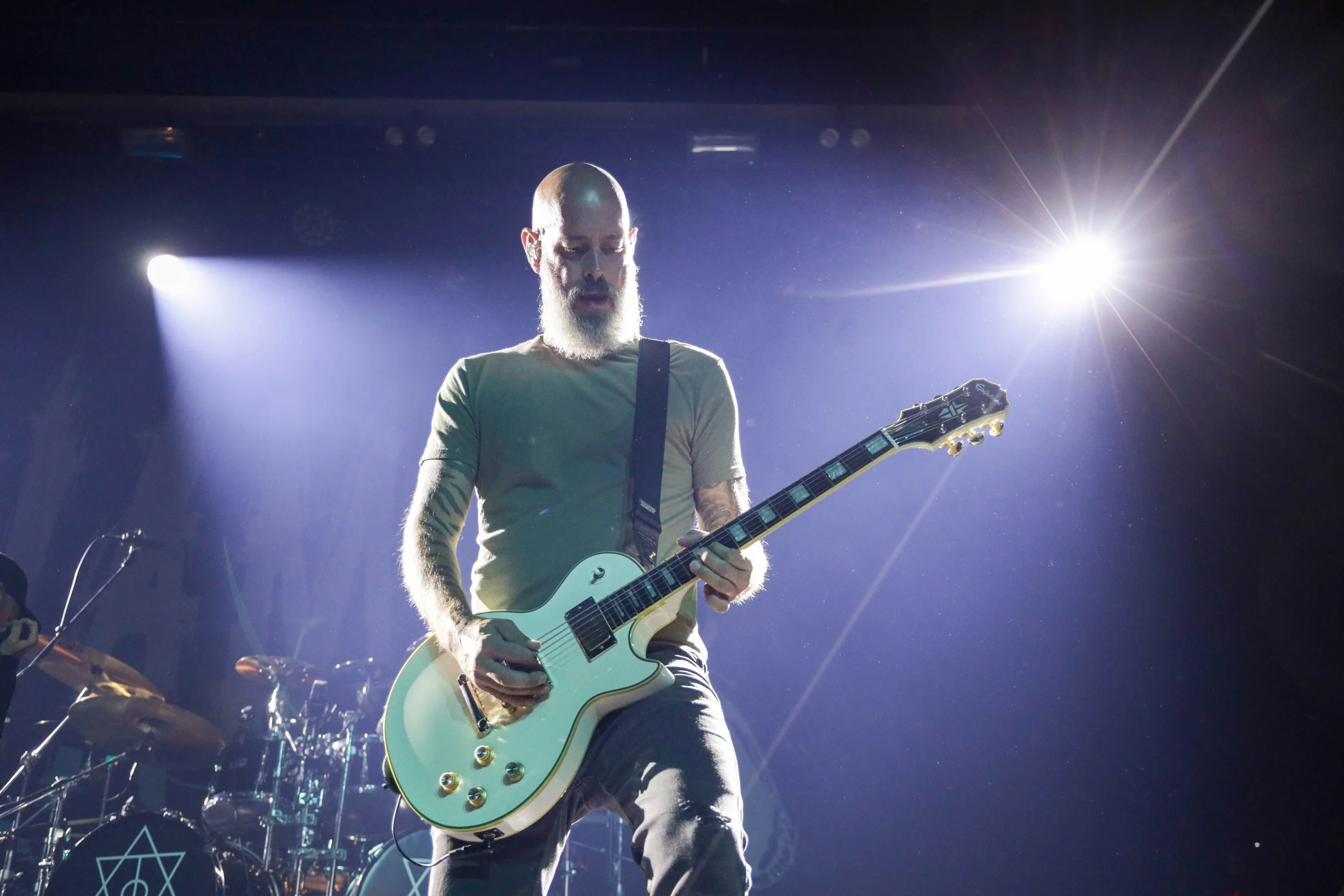 A bearded man playing an electric guitar on stage with bright stage lights shining behind him.