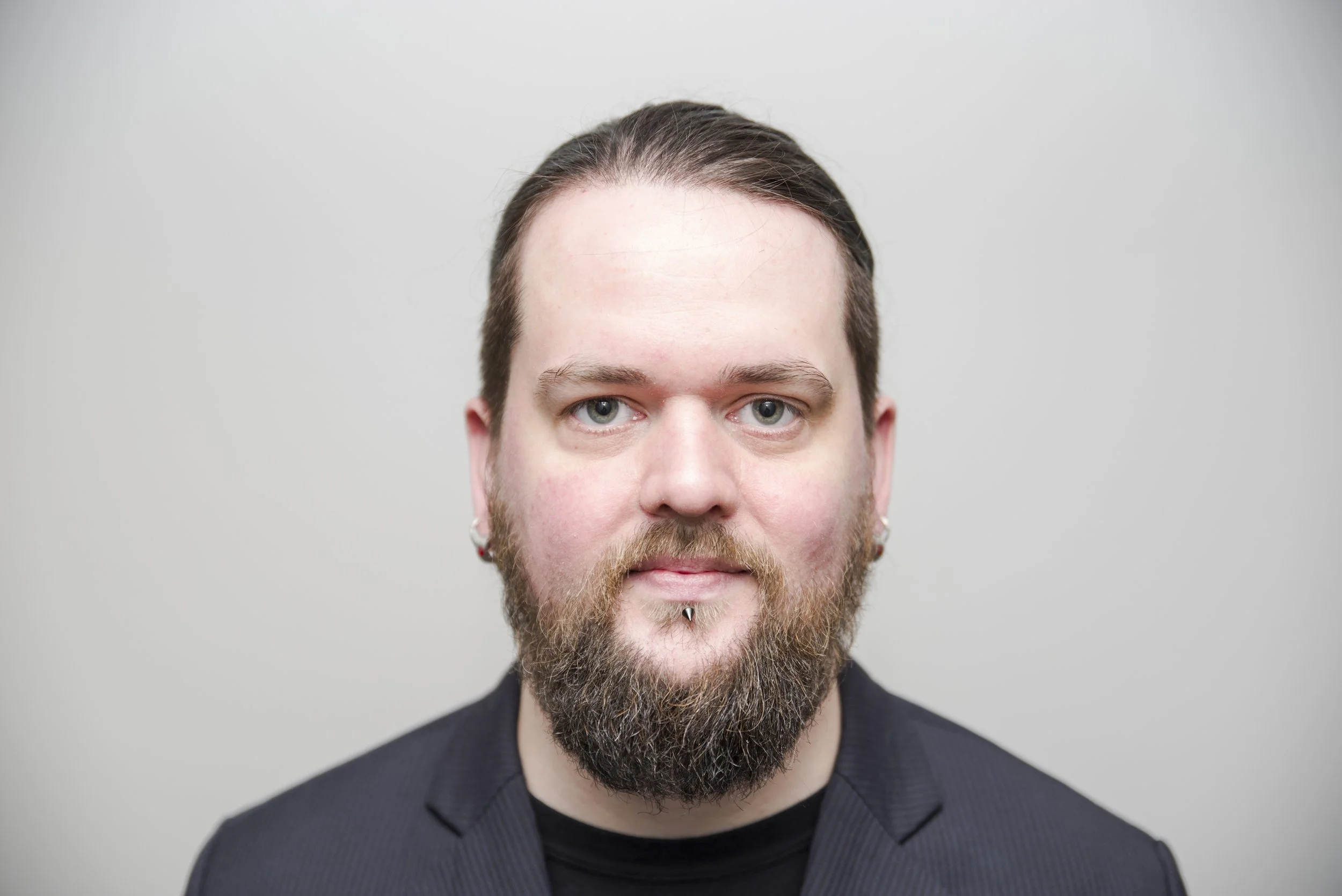 Close-up portrait of a man with a beard, piercings, and short hair, wearing a black blazer, against a plain light gray background.