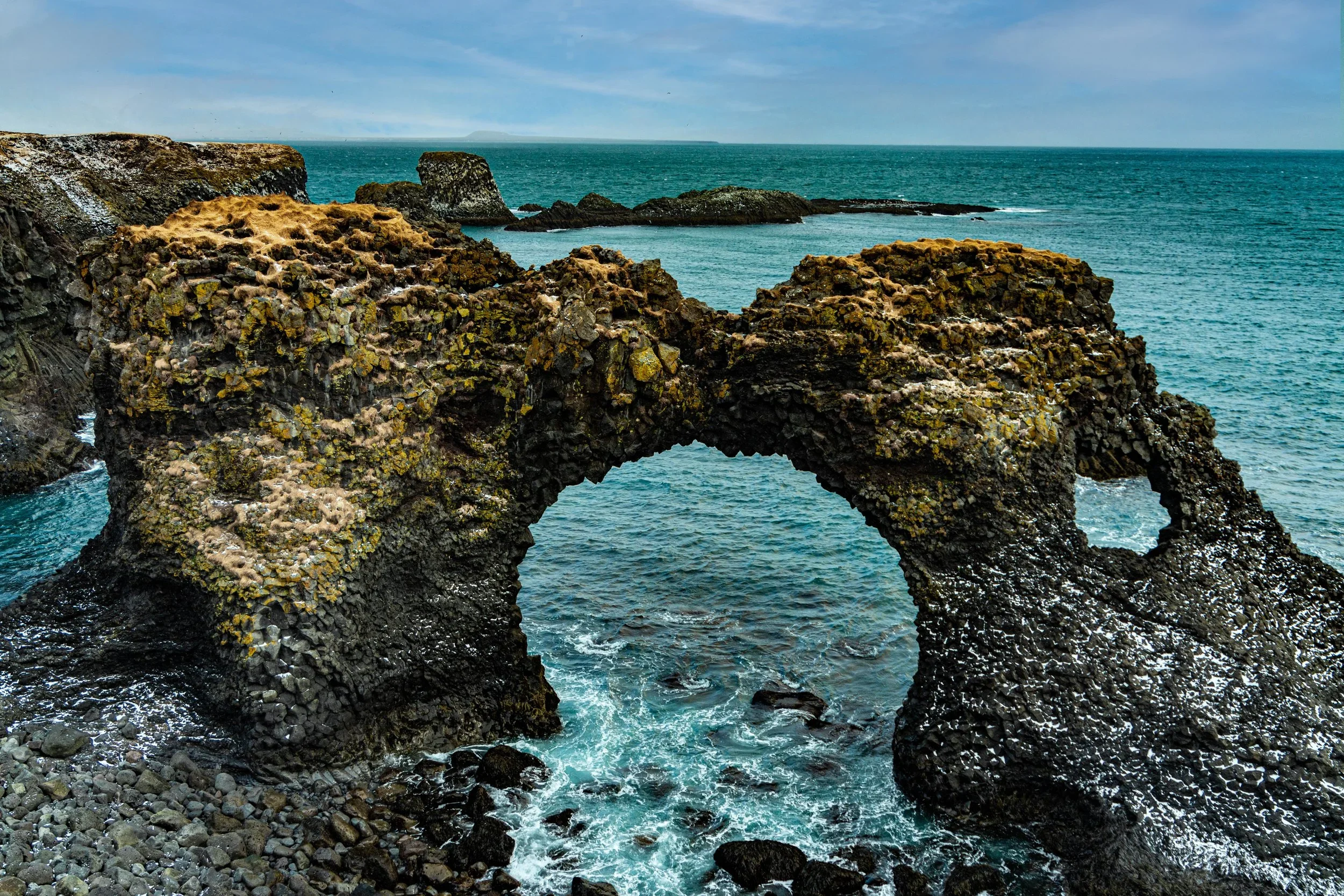 A natural rock formation on the ocean showing a large arch with rocky cliffs in the background and water crashing around the base.